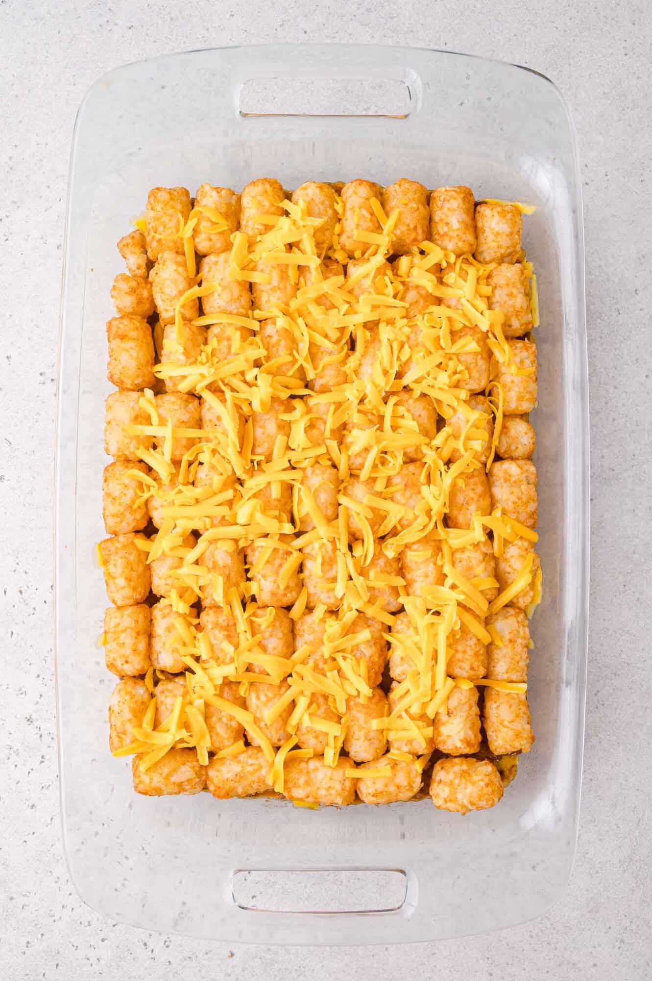 A glass baking dish filled with neatly arranged tater tots topped with shredded cheddar cheese on a light countertop.
