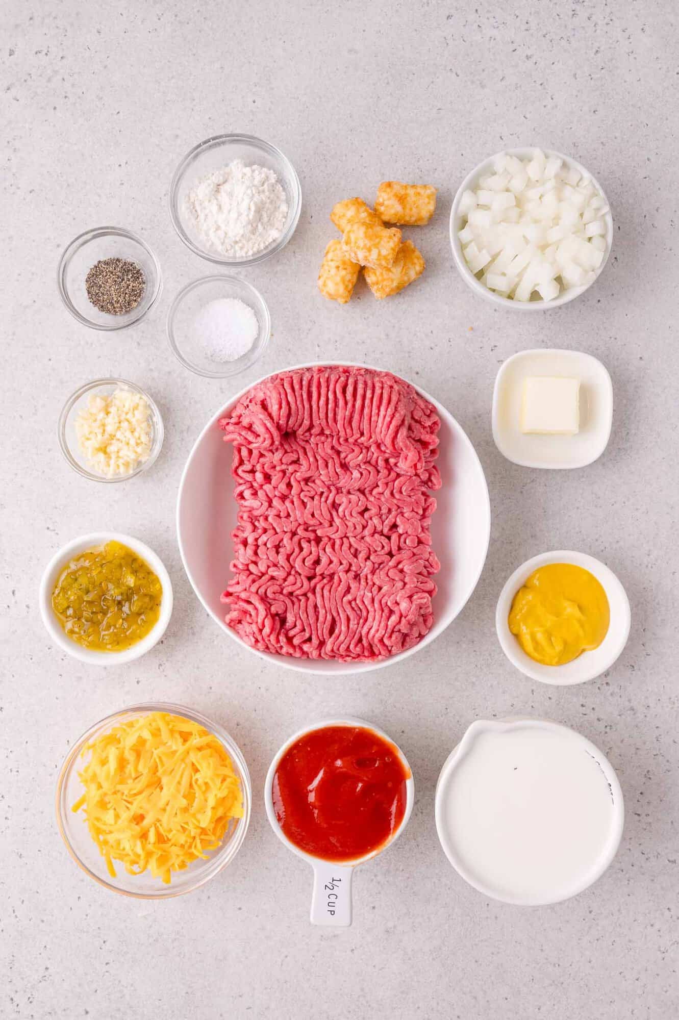 Ground beef, shredded cheese, diced onions, tater tots, milk, ketchup, mustard, relish, butter, flour, salt, pepper, and garlic arranged in bowls on a gray countertop.