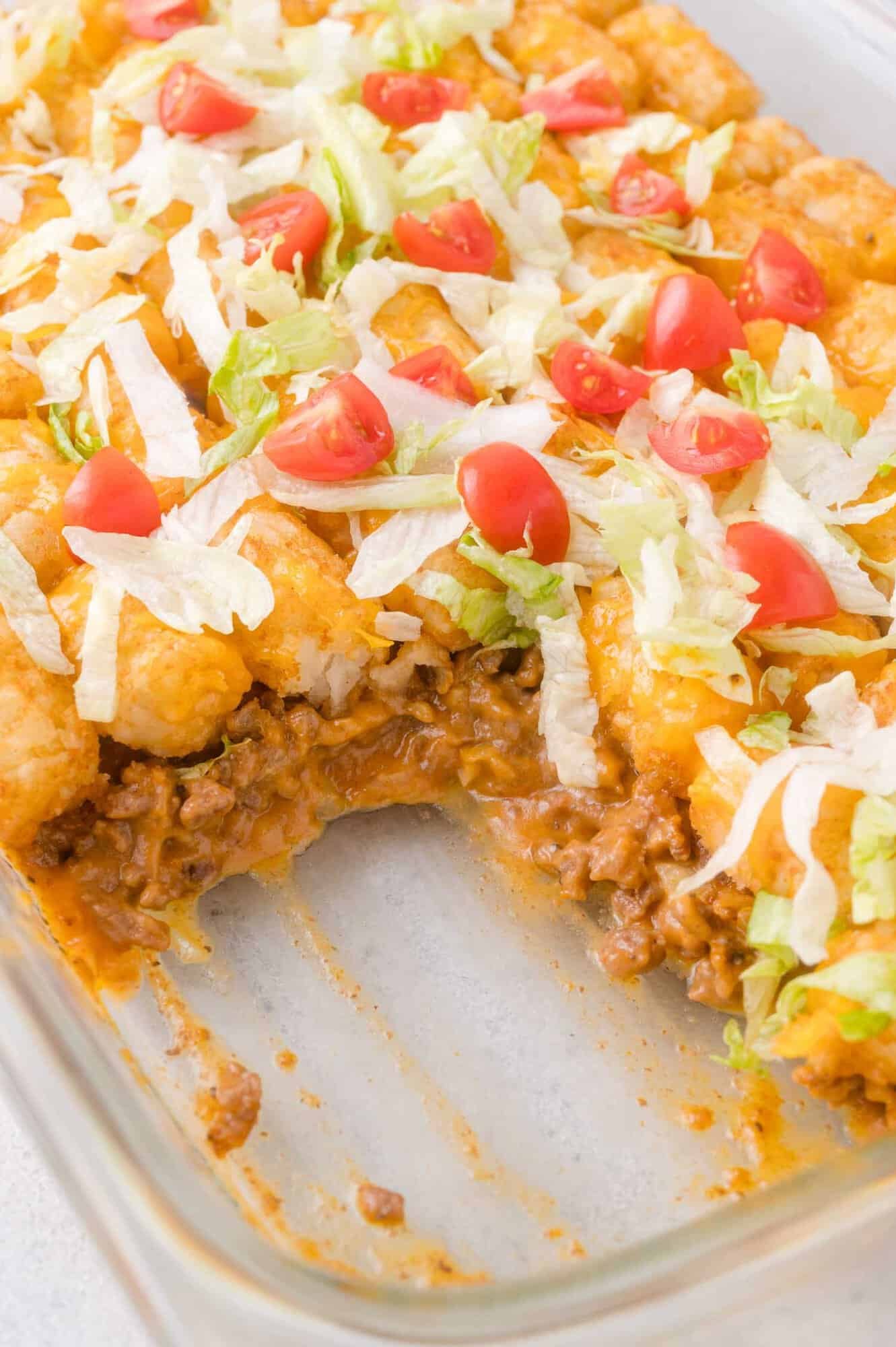 A casserole dish with a serving missing, showing layers of ground beef, cheese, tater tots, and toppings of shredded lettuce and sliced cherry tomatoes.