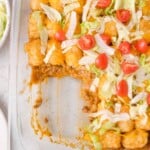 Glass baking dish with a portion of cheeseburger tater tot casserole topped with shredded lettuce, diced tomatoes, and melted cheese; one section is scooped out.