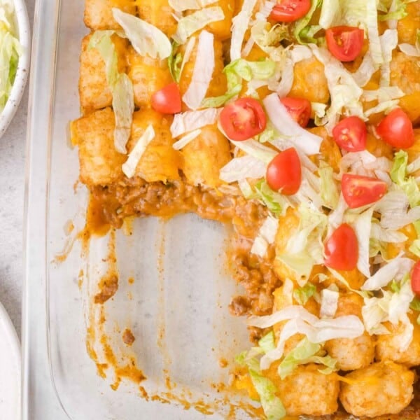 Glass baking dish with a portion of cheeseburger tater tot casserole topped with shredded lettuce, diced tomatoes, and melted cheese; one section is scooped out.