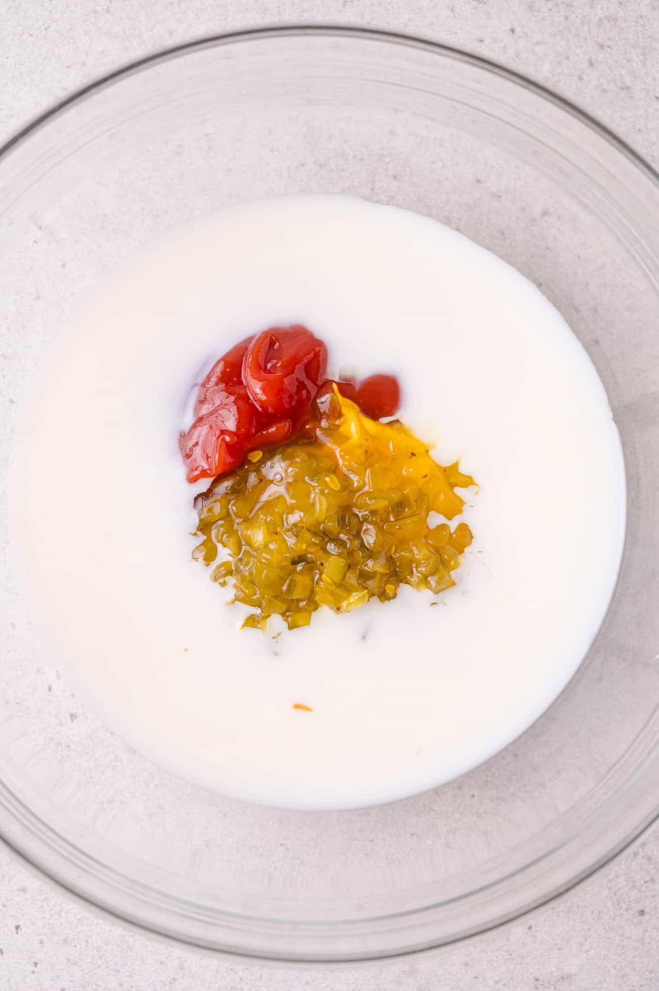 A glass bowl containing milk, ketchup, yellow mustard, and relish on a light surface.