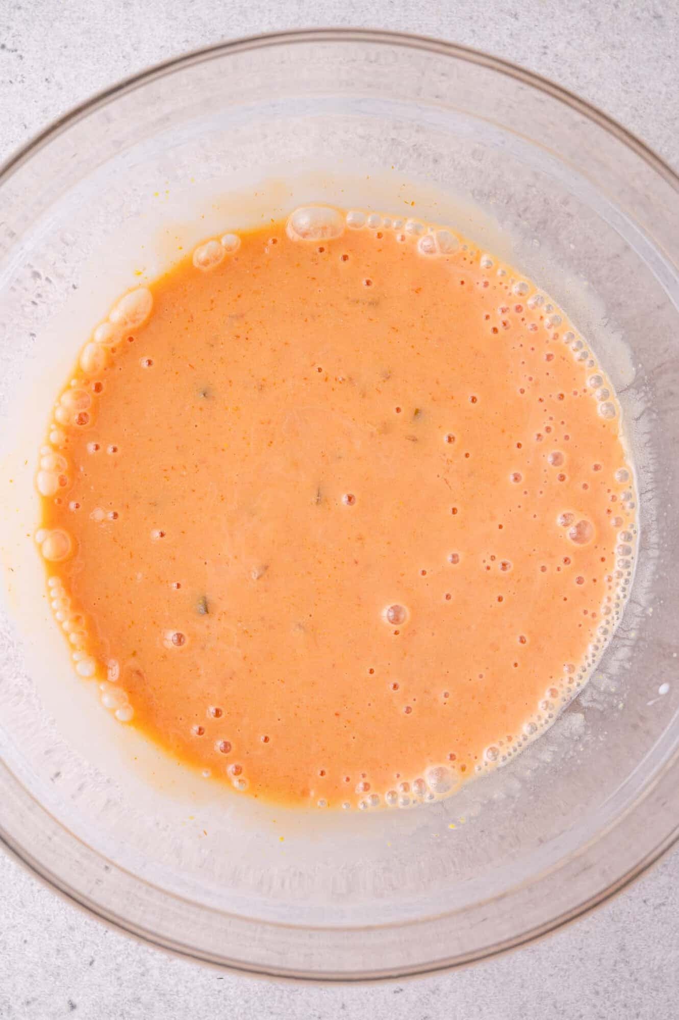 A clear glass bowl containing a frothy, orange mixture of the sauce ingredients with small bubbles on the surface, set on a light-colored countertop.