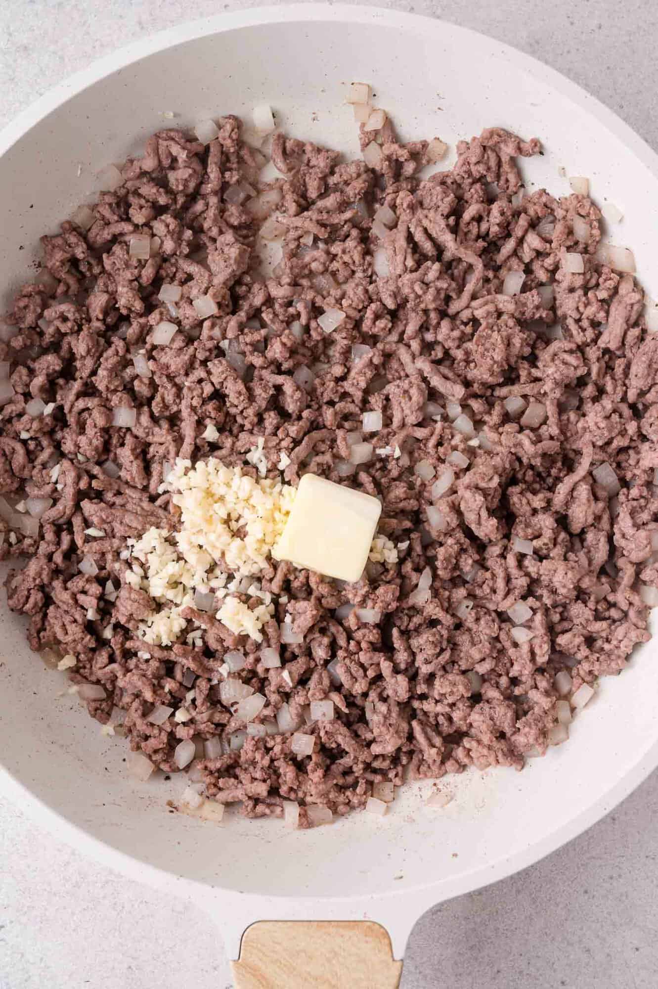 Ground beef and diced onions are browning in a skillet, with chopped garlic and a pat of butter added in the center.