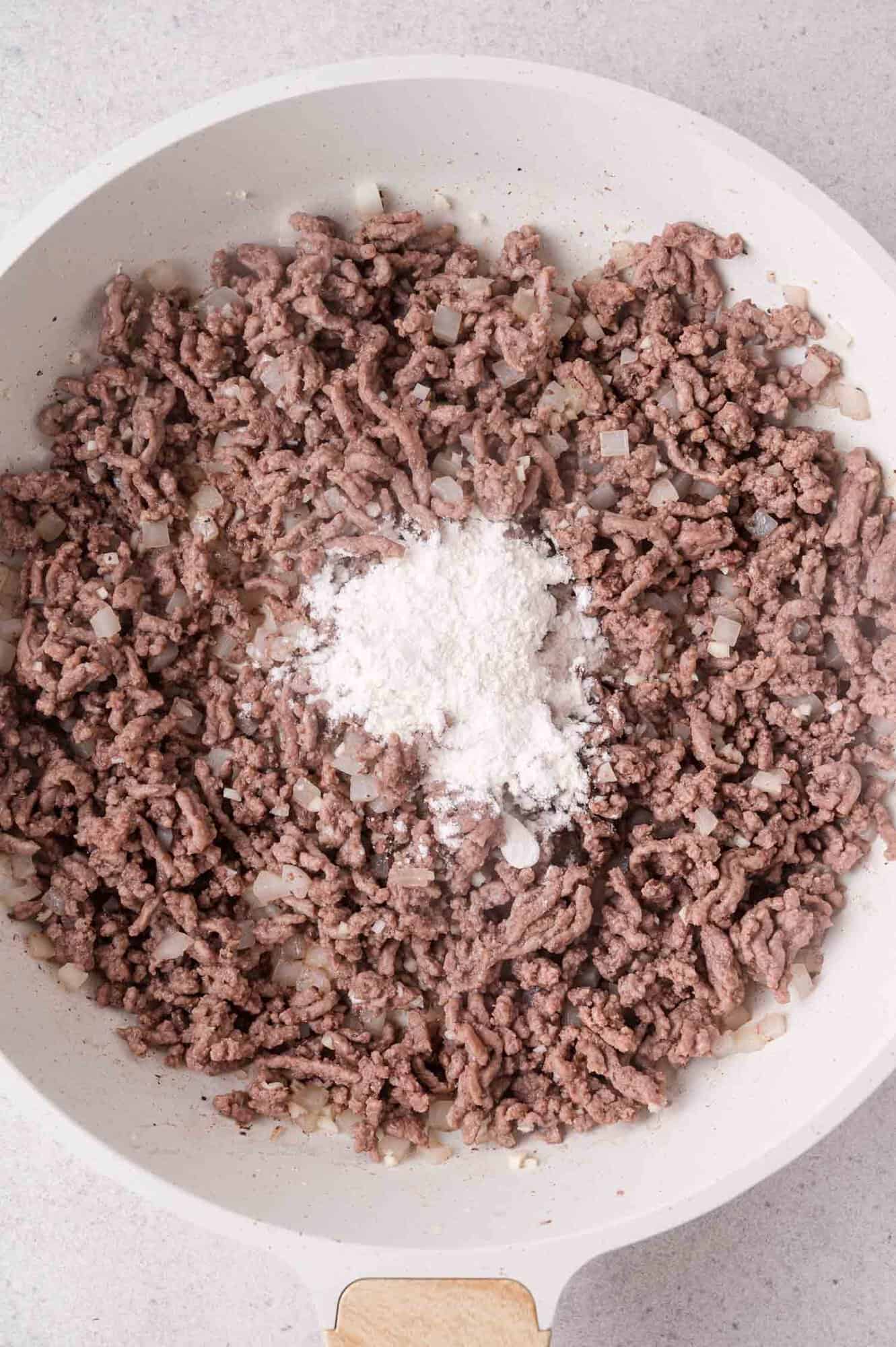 Ground beef cooking in a white skillet with diced onions and a mound of white flour added on top.