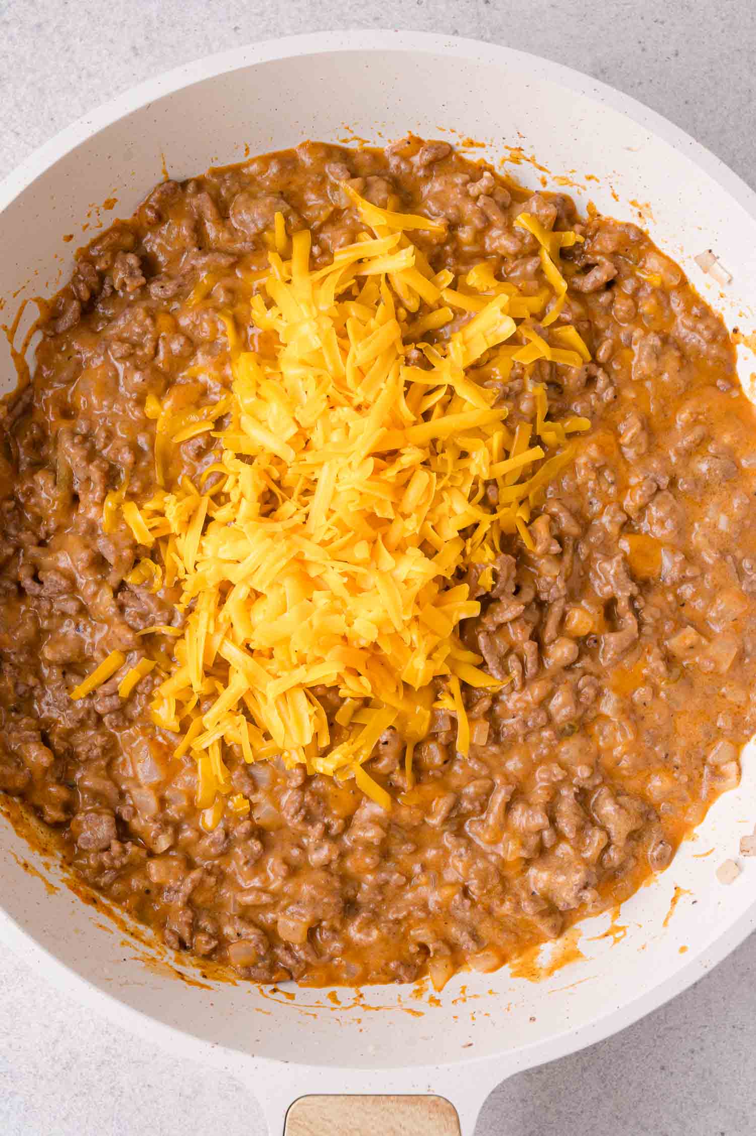 Ground beef in a creamy sauce is cooking in a white skillet, topped with a pile of shredded cheddar cheese.