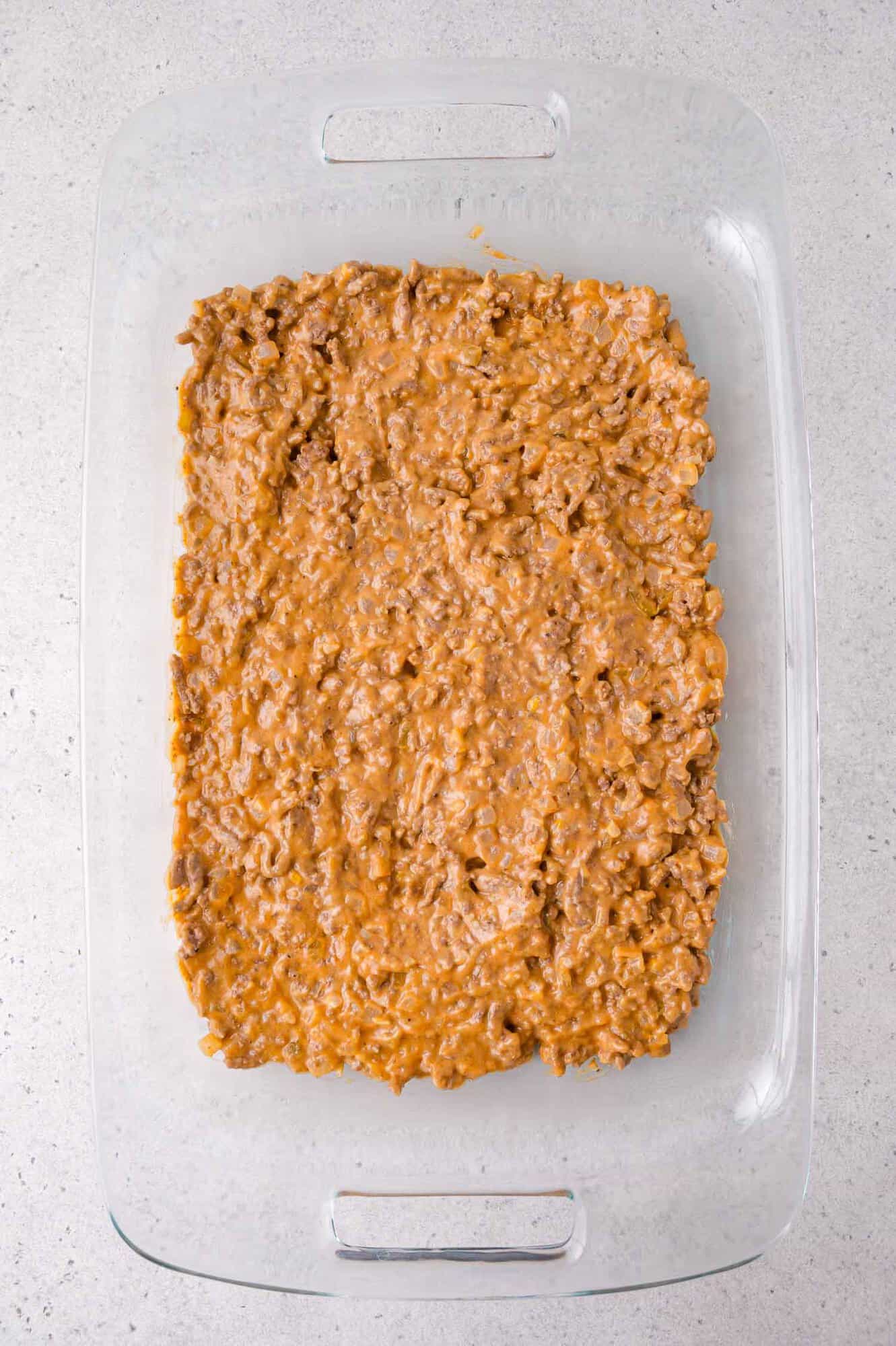A glass baking dish filled with an even layer of ground beef mixed with a creamy sauce, placed on a light gray countertop.