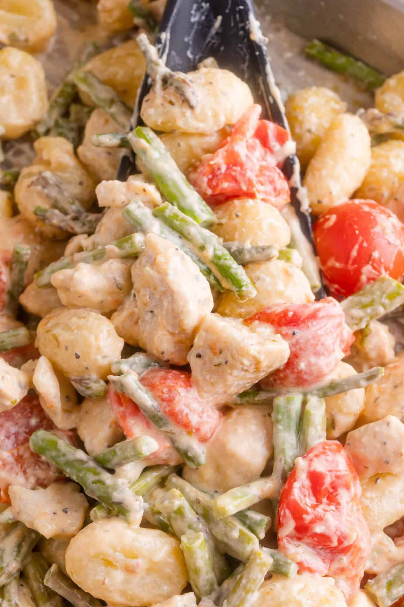 Close-up of creamy gnocchi with chicken, asparagus, and cherry tomatoes, being stirred with a black spoon.