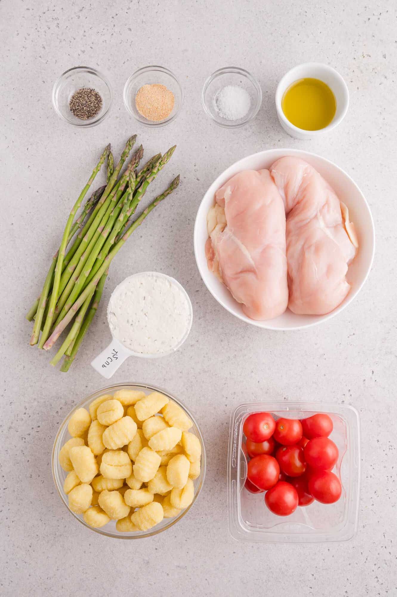 Raw chicken breasts, asparagus, gnocchi, cherry tomatoes, Parmesan cheese, olive oil, salt, pepper, and garlic powder are arranged in separate bowls on a light countertop.