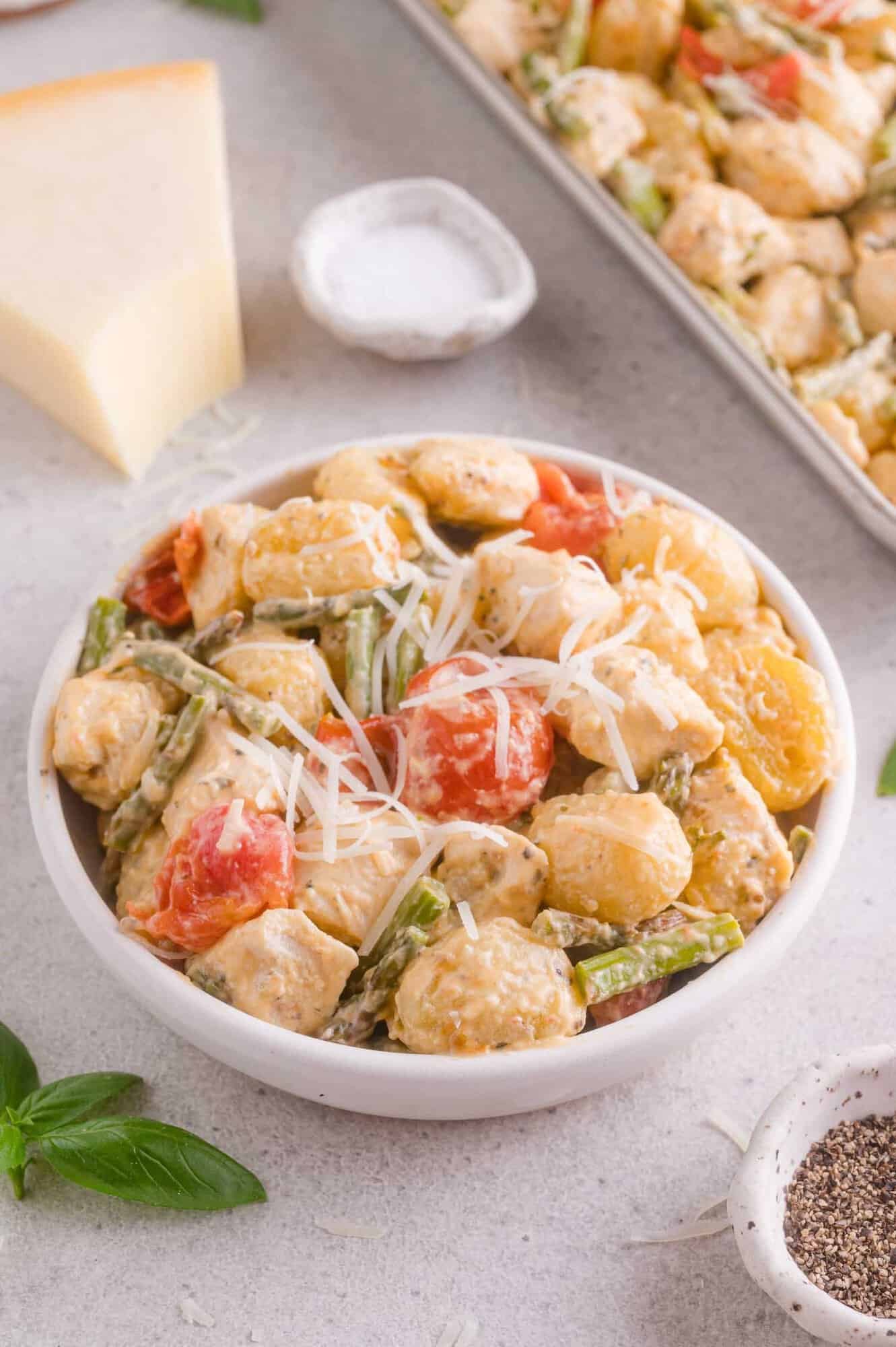 Bowl of creamy gnocchi with chicken, asparagus, tomatoes, and shredded cheese, with salt, pepper, cheese, and basil visible in the background.