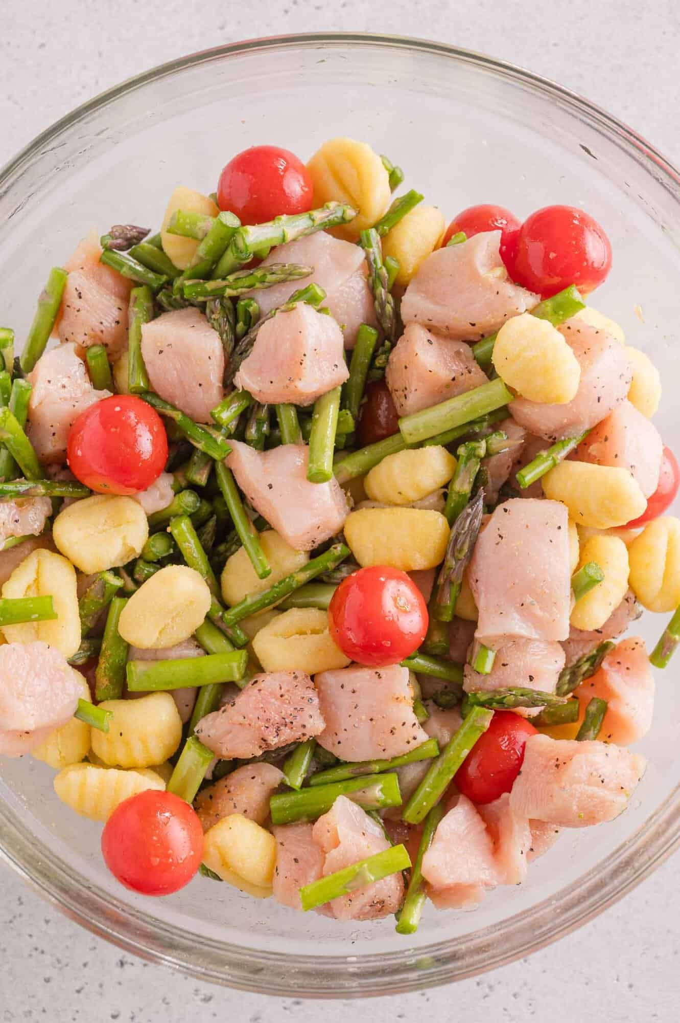 A glass bowl contains raw diced chicken, gnocchi, cherry tomatoes, and chopped asparagus, all seasoned with black pepper.