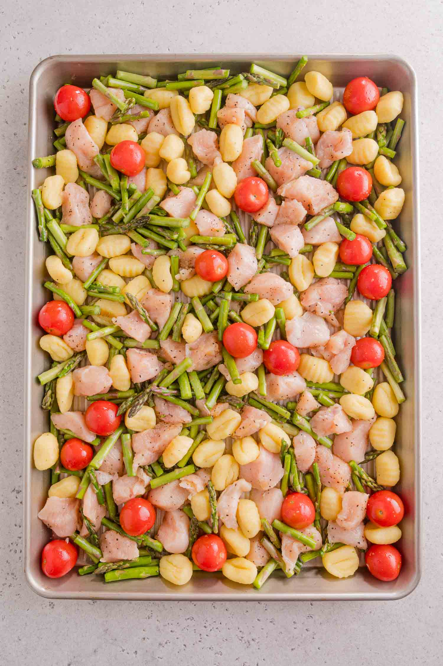 A sheet pan with uncooked diced chicken, gnocchi, asparagus, and cherry tomatoes, all evenly spread out and seasoned, ready to bake.
