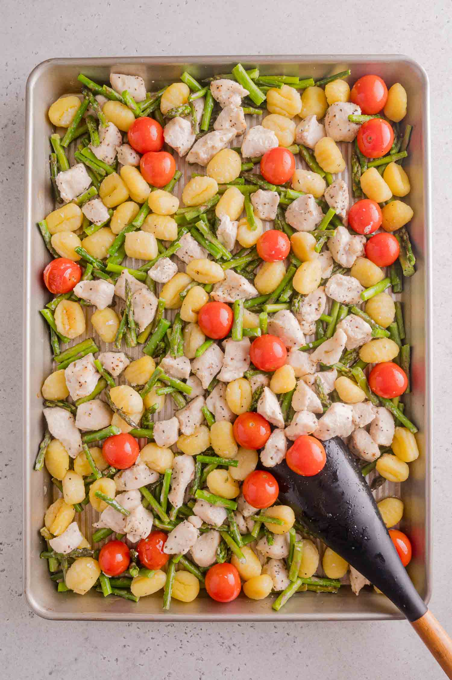 Sheet pan with cooked gnocchi, diced chicken, asparagus pieces, and cherry tomatoes, mixed with a black spatula on a light countertop.