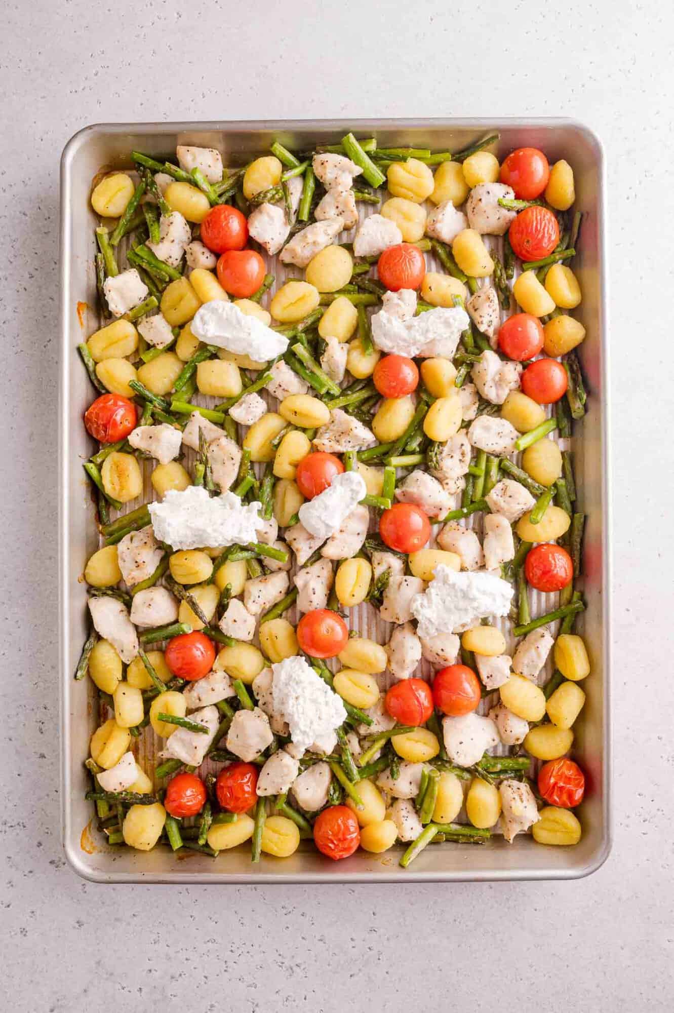 A baking sheet with gnocchi, cherry tomatoes, asparagus, diced chicken, and dollops of cream cheese cheese on a light countertop.