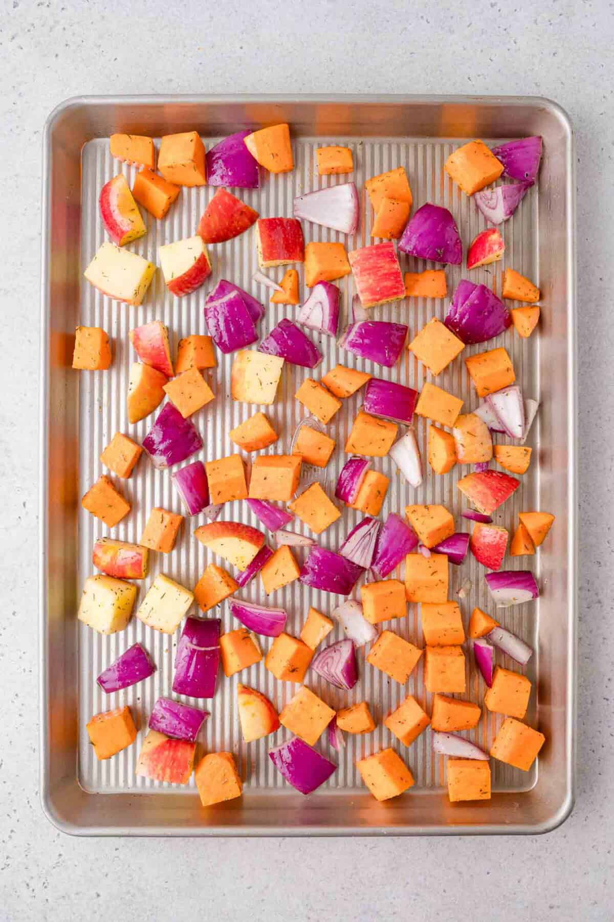 Diced sweet potatoes, red onions, and apples spread evenly on a metal baking sheet, ready for roasting.