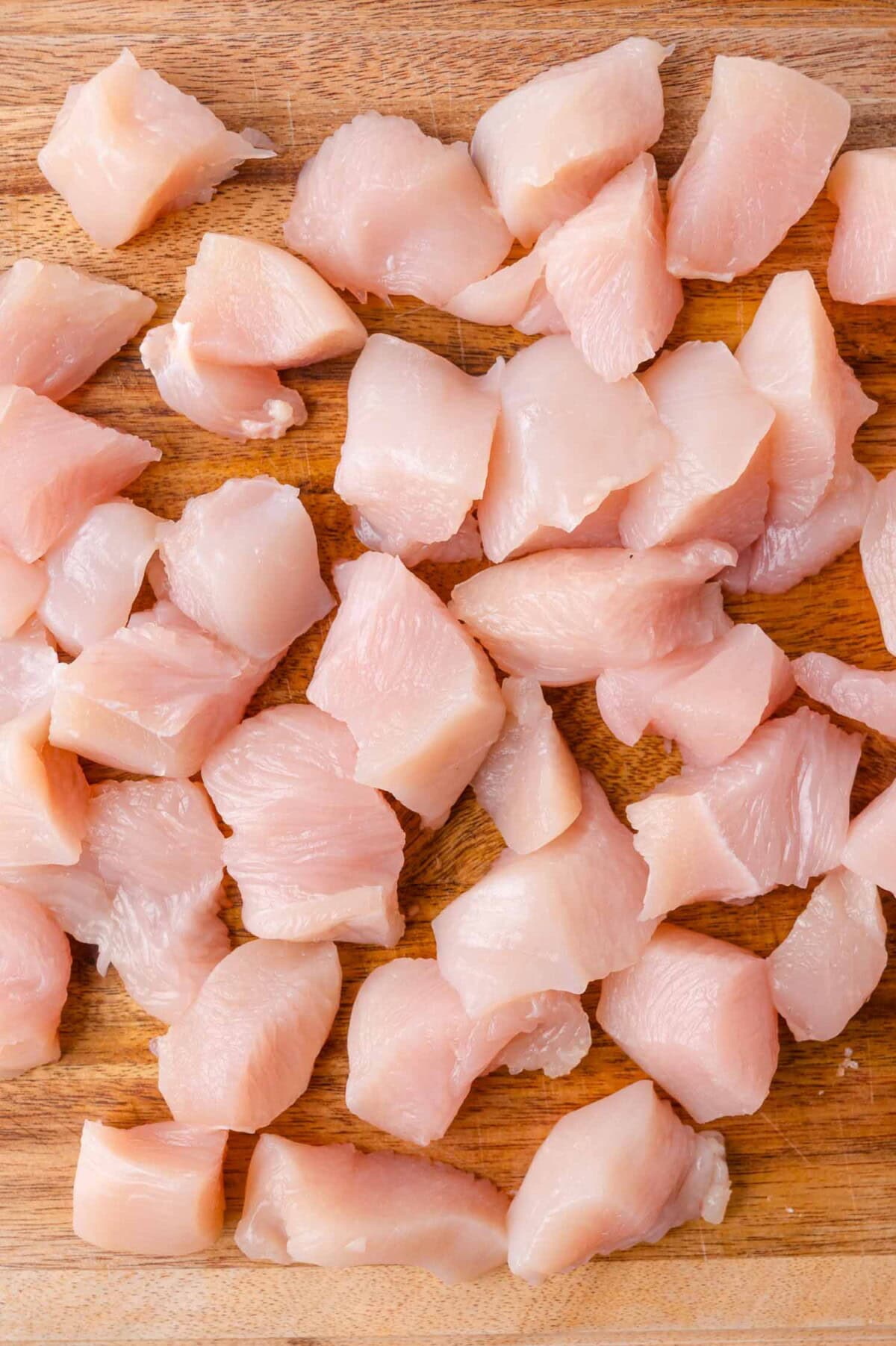 Raw, cubed pieces of chicken breast are spread out on a wooden cutting board.