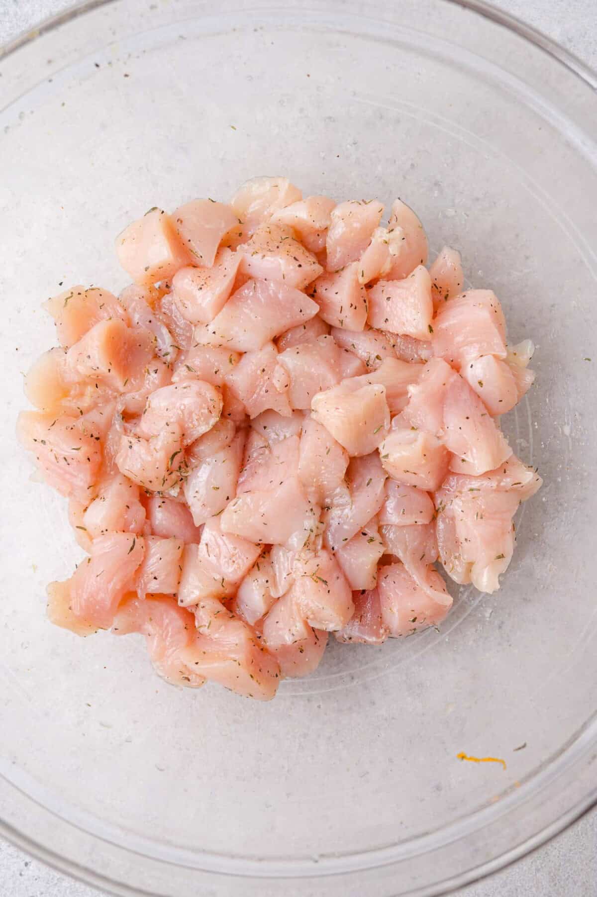 Diced raw chicken pieces seasoned with herbs in a clear mixing bowl, viewed from above.