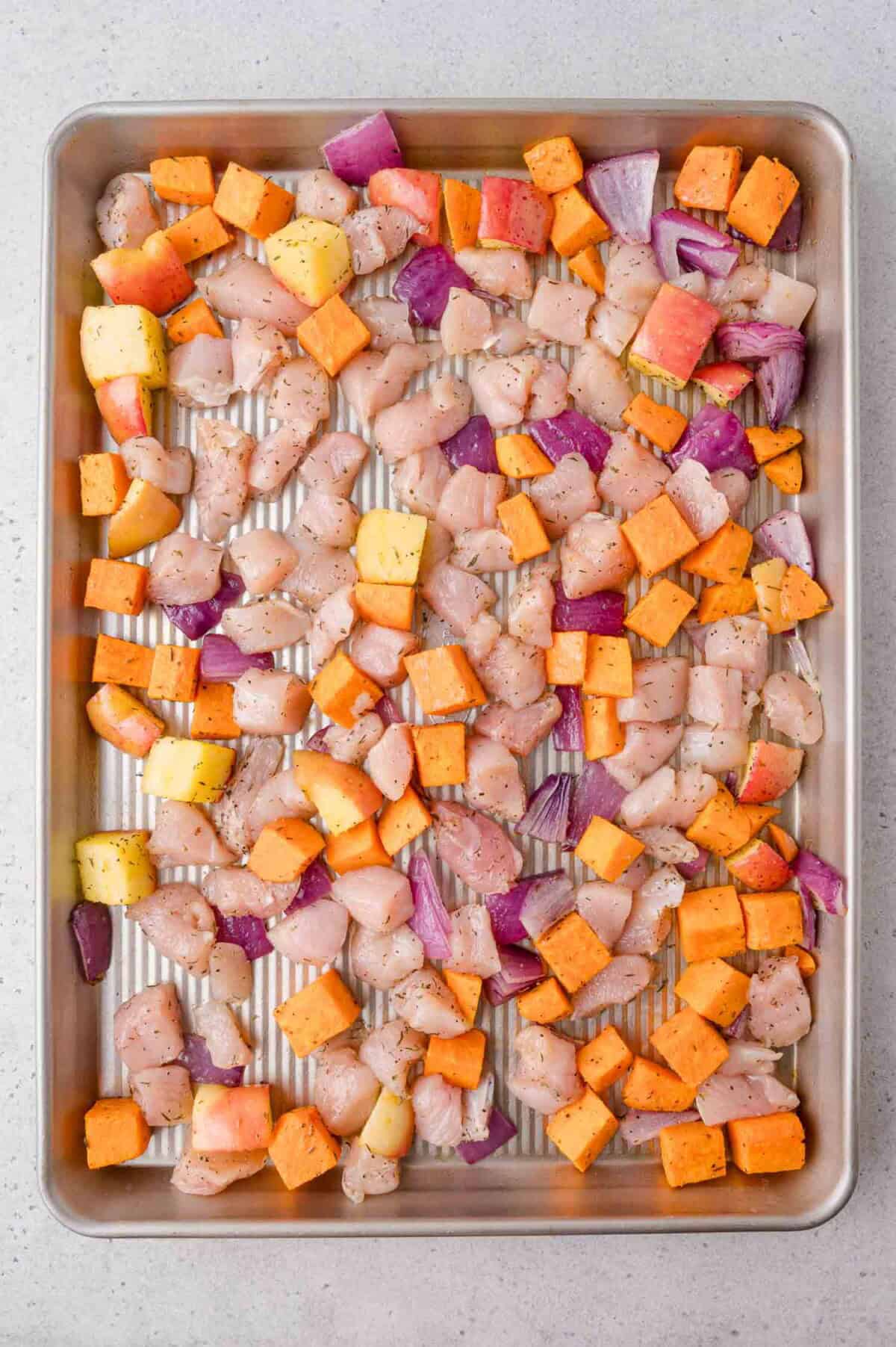 Diced chicken, sweet potatoes, apples, and red onions are spread evenly on a baking sheet, ready for roasting.