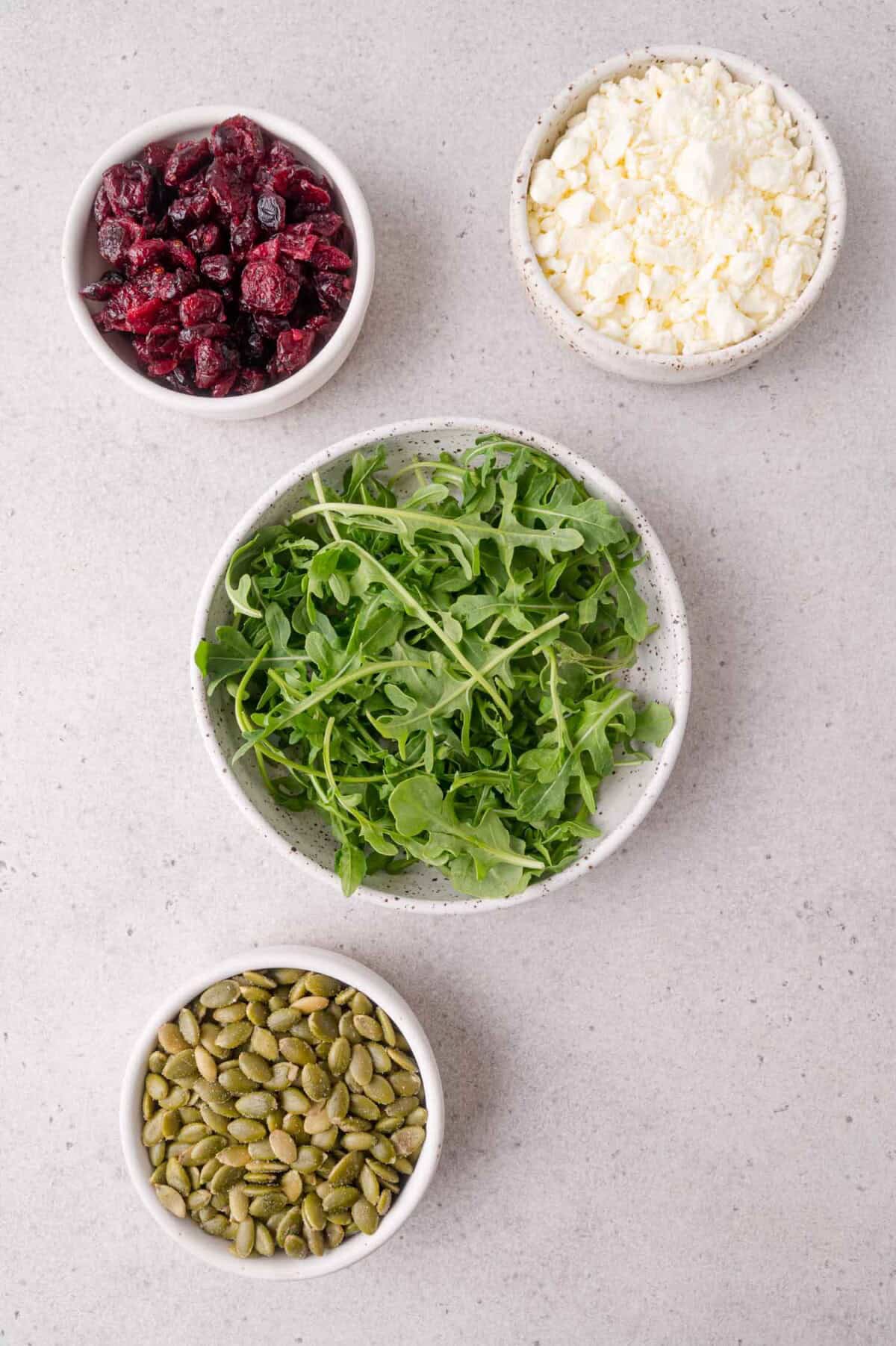 Four bowls containing dried cranberries, crumbled feta cheese, arugula leaves, and pumpkin seeds are arranged on a light grey surface.