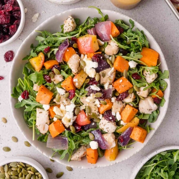 A bowl of salad with arugula, roasted sweet potatoes, red onions, chicken, cranberries, feta cheese, and pumpkin seeds, surrounded by small bowls of salad ingredients.