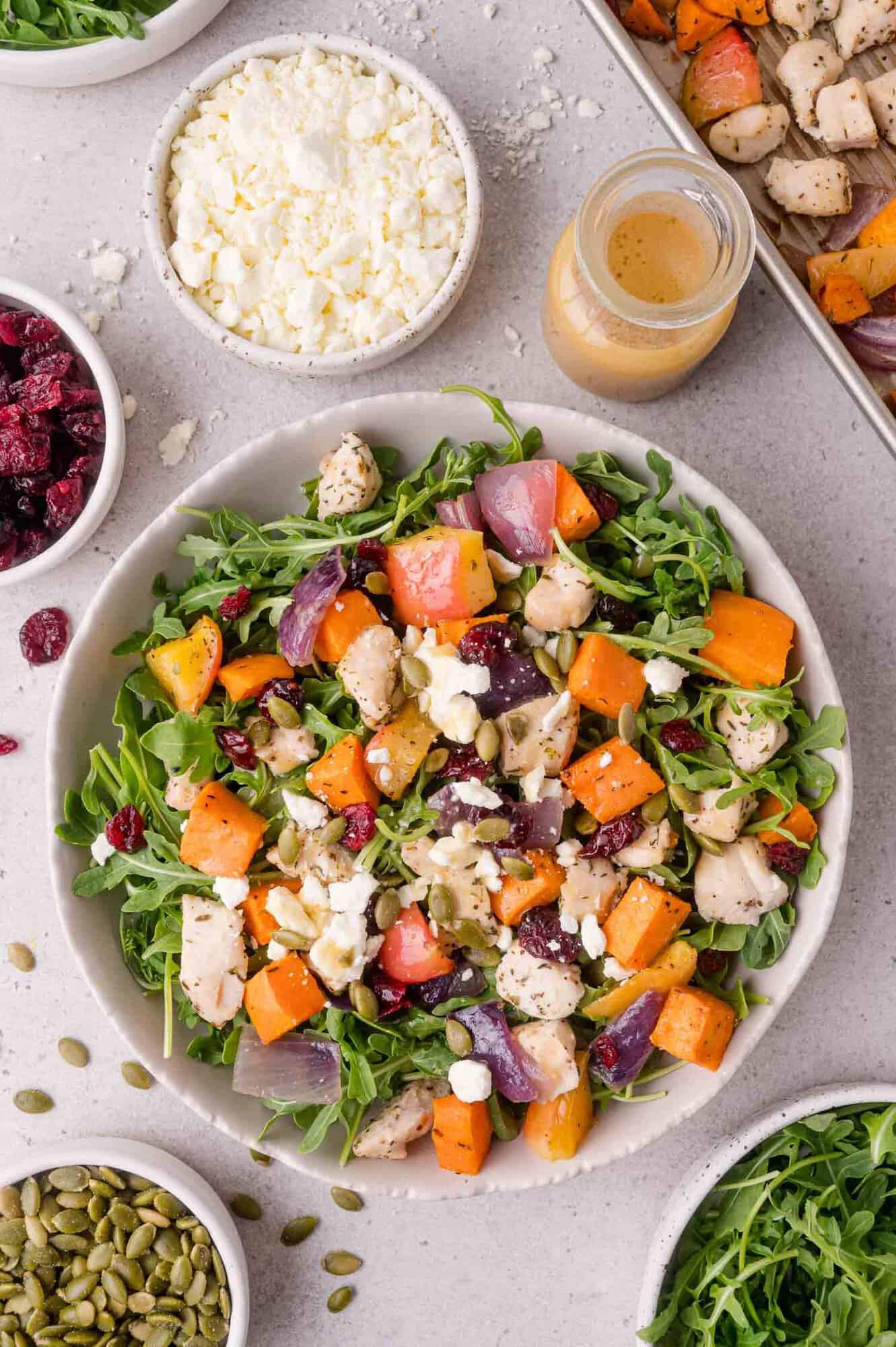 A bowl of salad with arugula, roasted sweet potatoes, chicken, red onions, dried cranberries, feta cheese, and seeds. Surrounding bowls hold ingredients and a small bottle of dressing.