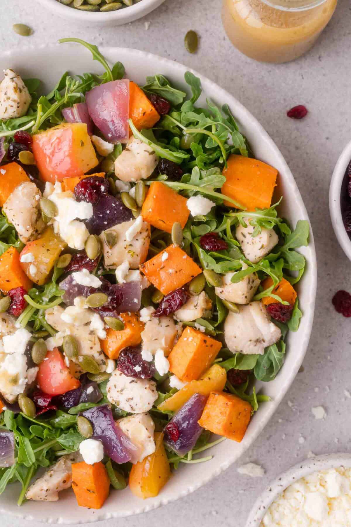 A salad with arugula, roasted sweet potatoes, red onion, chicken, feta cheese, pumpkin seeds, and dried cranberries in a white bowl, with small bowls and dressing nearby.