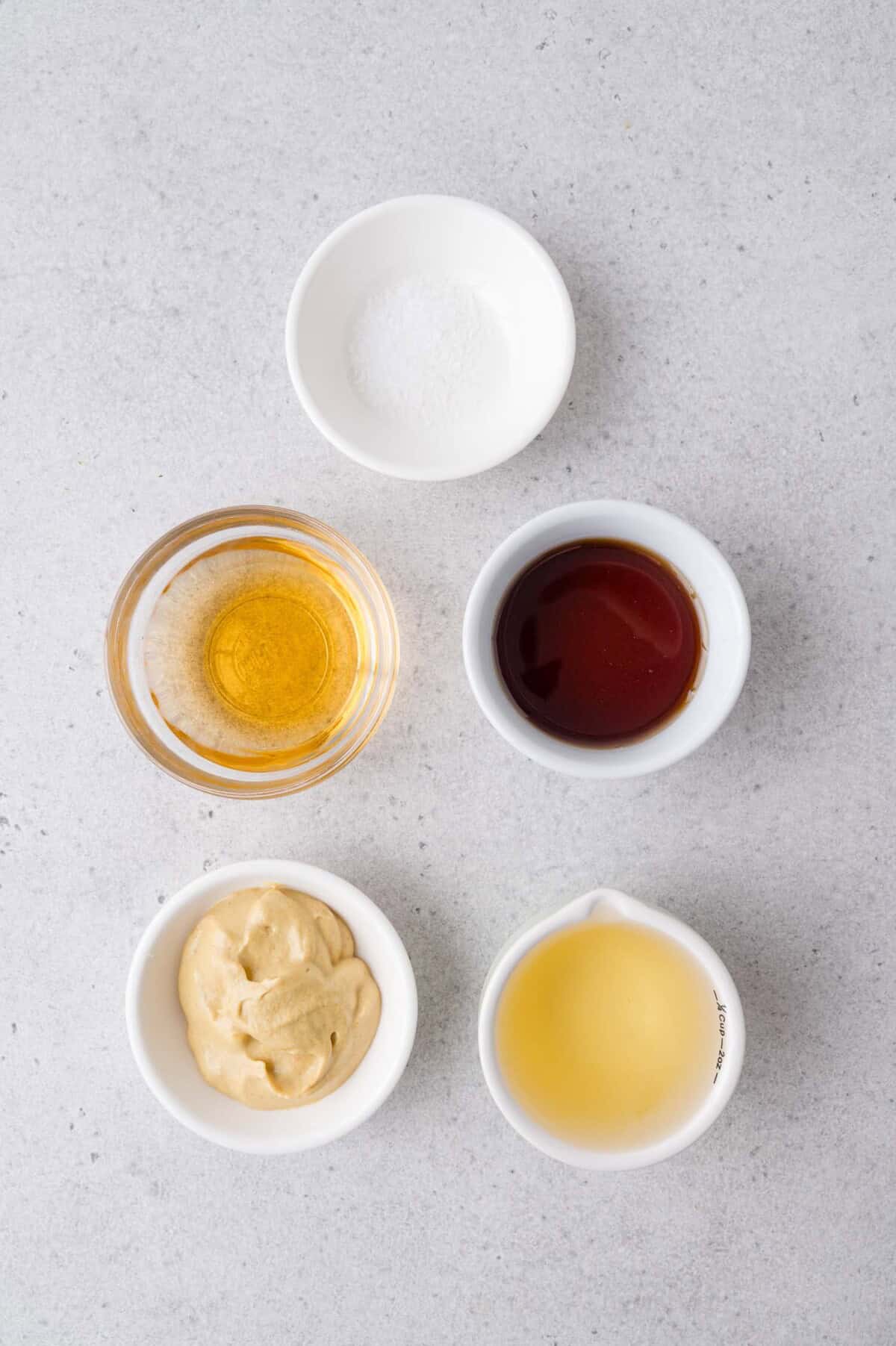 Five small bowls containing salt, olive oil, mustard, apple cider vinegar, and maple syrup, arranged on a light gray surface.