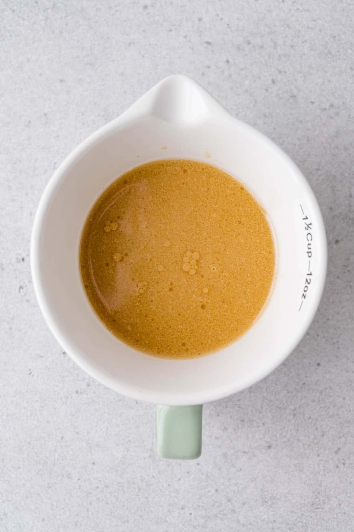 White ceramic measuring cup with a green handle containing a light brown dressing, viewed from above, on a light gray surface.