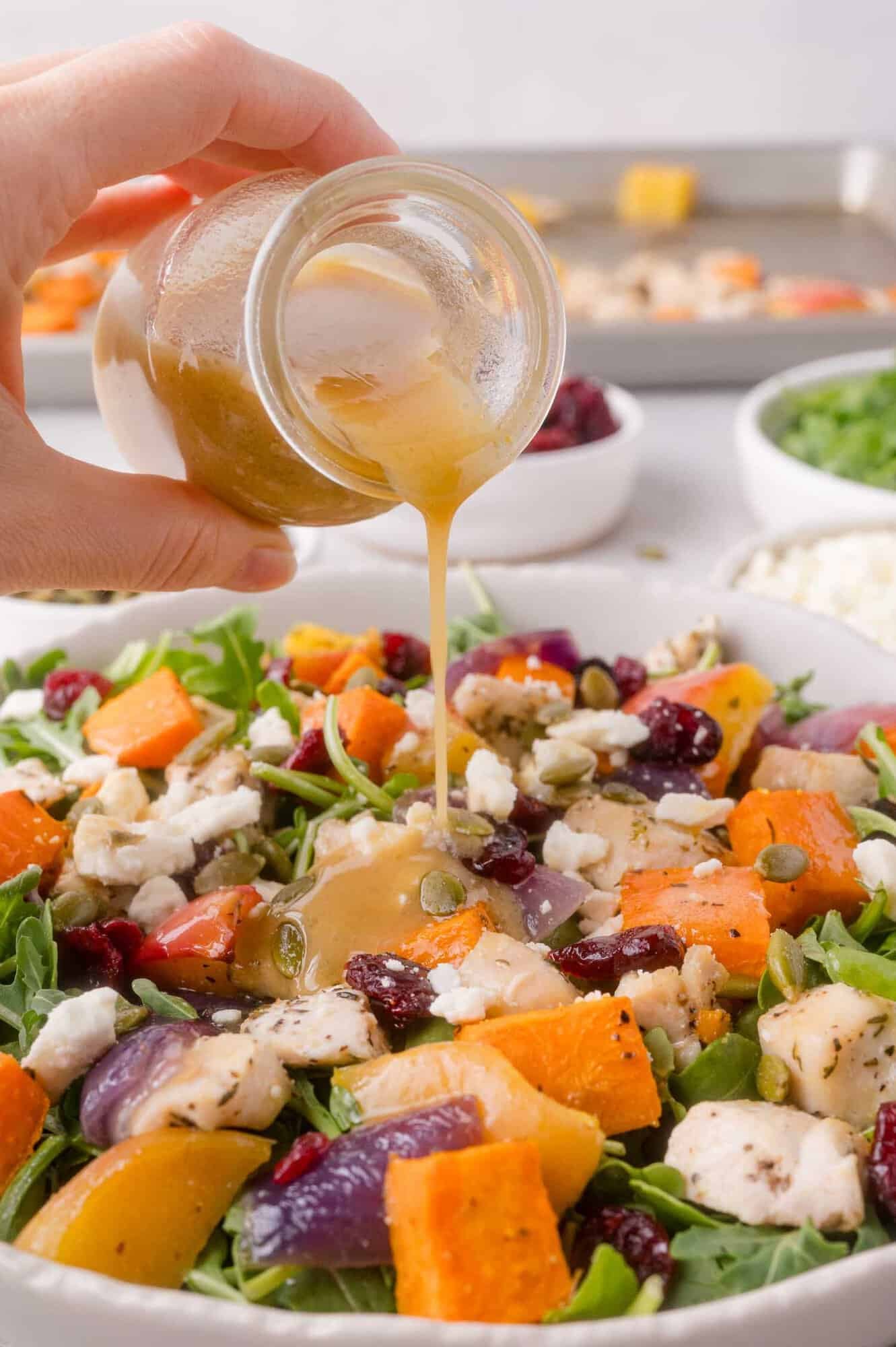 A hand pours dressing over a salad with roasted sweet potatoes, red onions, greens, cranberries, seeds, and cheese.
