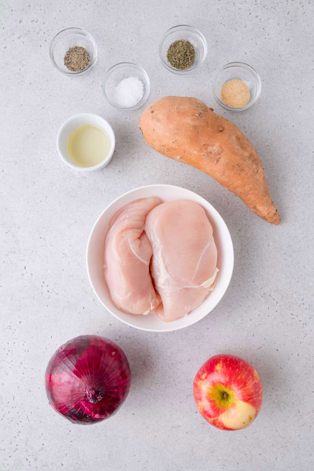 Raw chicken breasts in a bowl, sweet potato, red onion, apple, oil, salt, pepper, and garlic powder arranged on a light gray surface.
