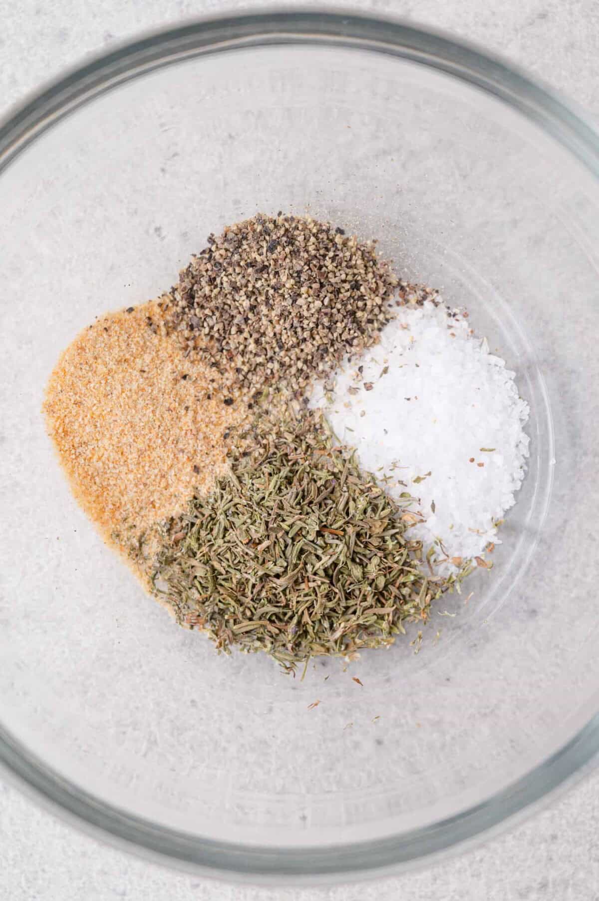 A glass bowl containing four piles of spices: ground black pepper, coarse salt, dried thyme, and garlic powder, on a light countertop.