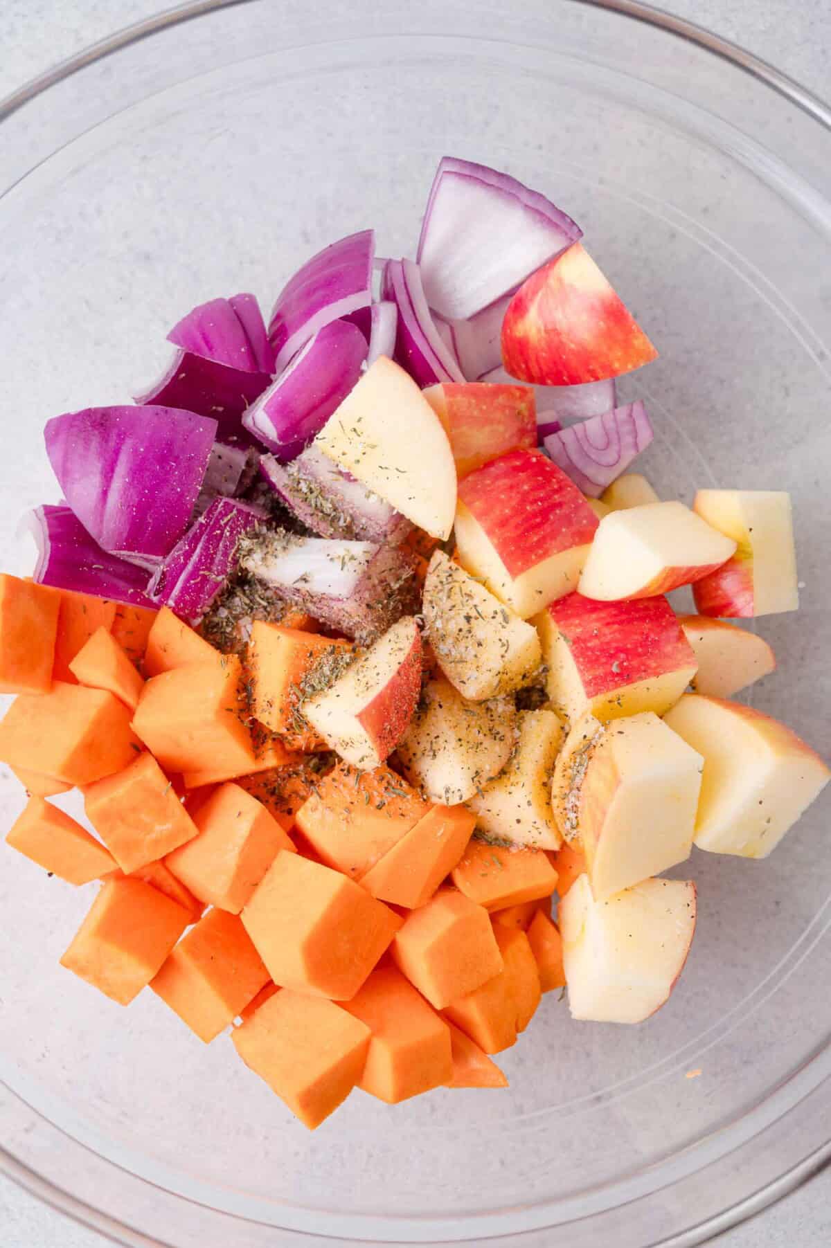 Chopped sweet potatoes, red onions, and apples with dried herbs in a clear glass bowl.