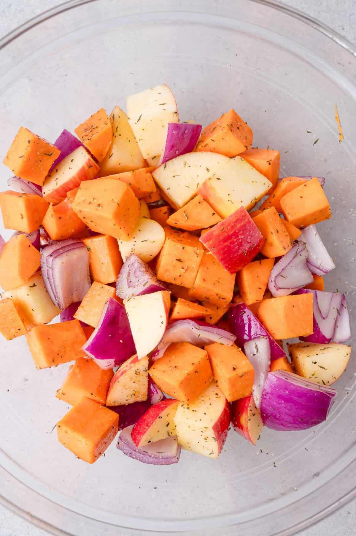 A clear bowl contains chopped sweet potatoes, red onions, and apples, all seasoned with herbs.