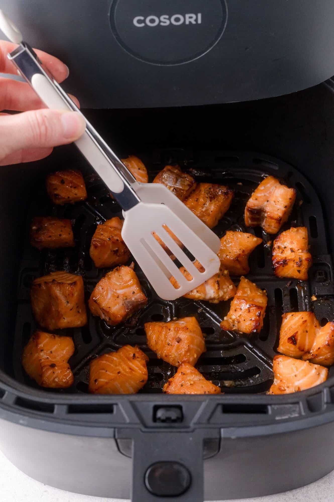 Hand holding a spatula above cooked salmon bites inside an open Cosori air fryer basket.