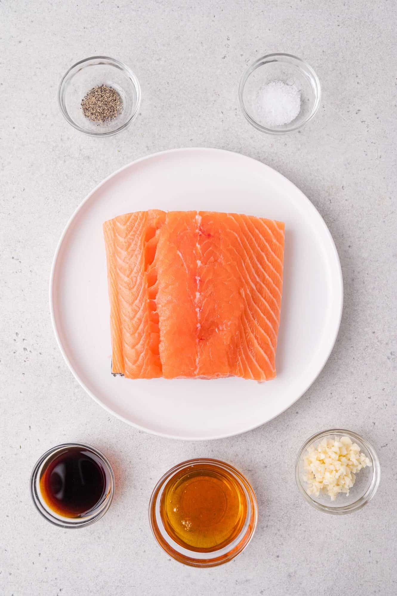 Raw salmon fillets on a white plate, surrounded by bowls of soy sauce, honey, minced garlic, black pepper, and salt on a light gray surface.
