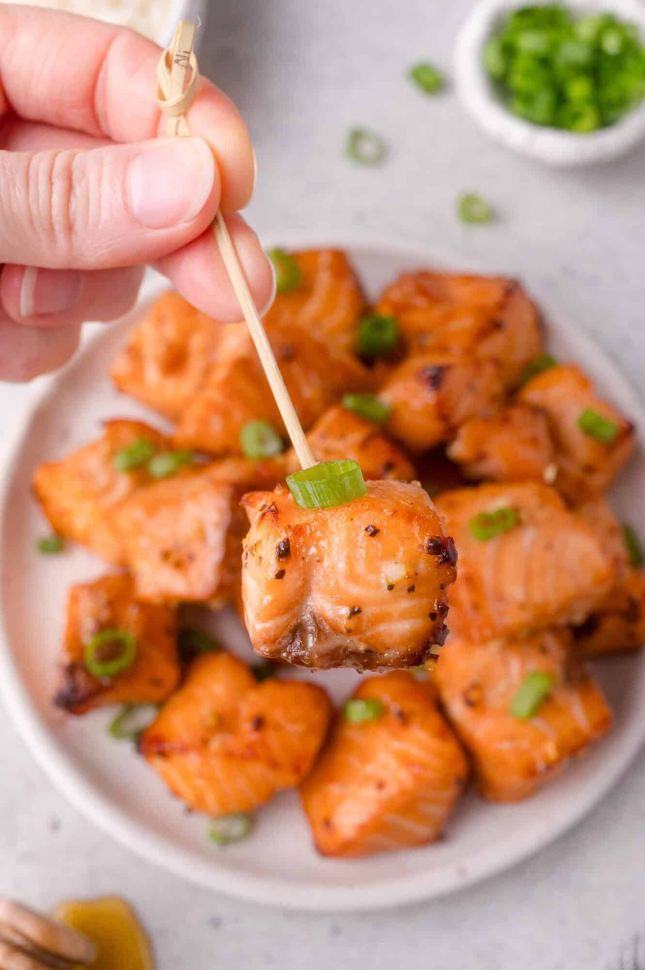 A hand holds a bamboo skewer with a glazed salmon bite, garnished with green onion, above a plate of similar salmon bites.
