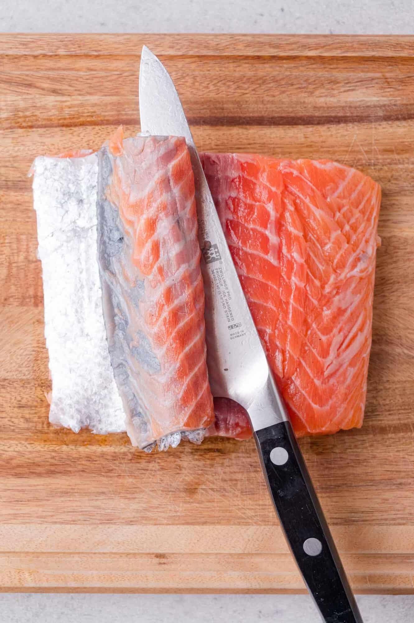 A knife is being used to separate the skin from a raw salmon fillet on a wooden cutting board.