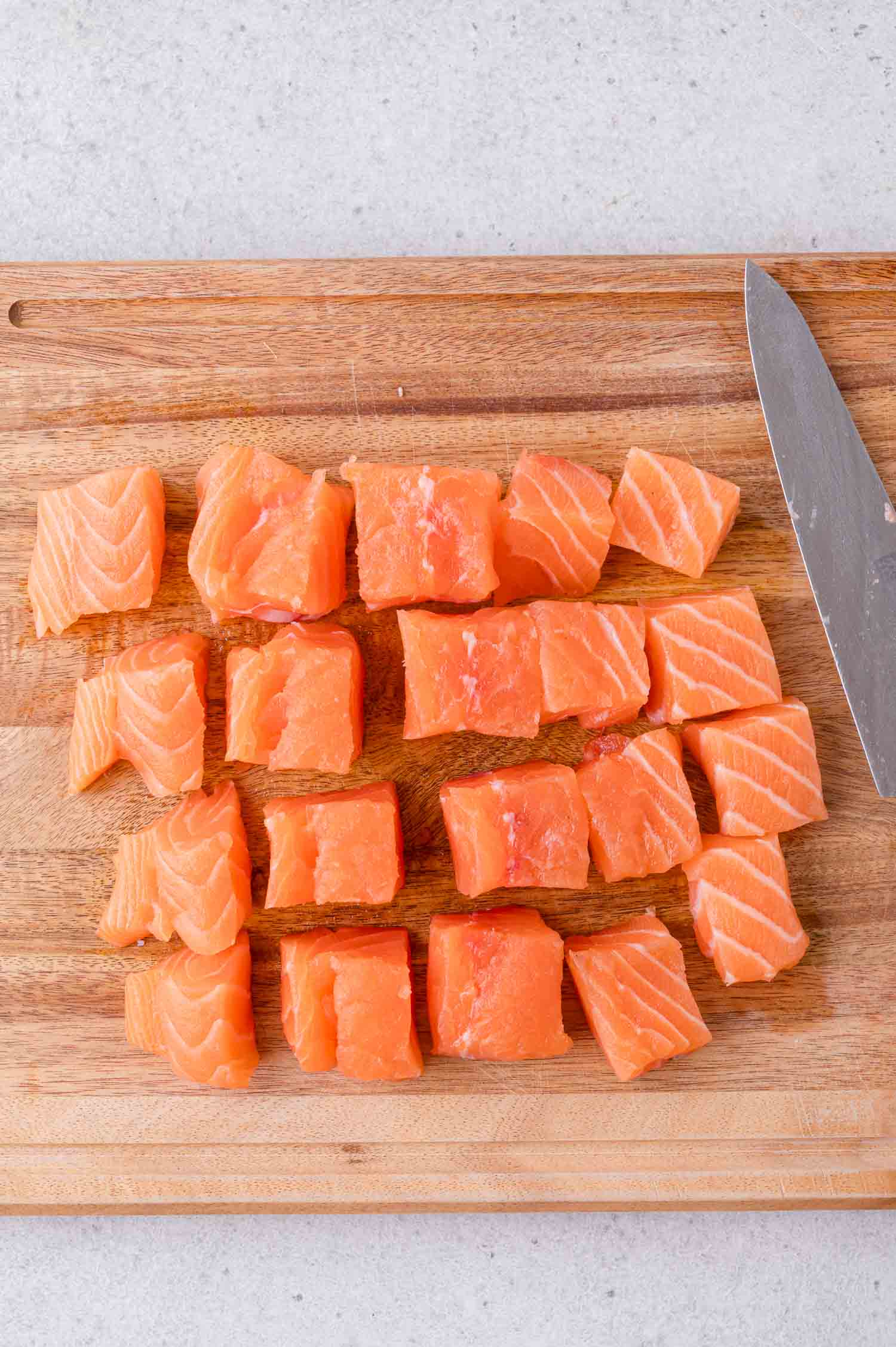 Cubed pieces of raw salmon are arranged in rows on a wooden cutting board next to a knife.