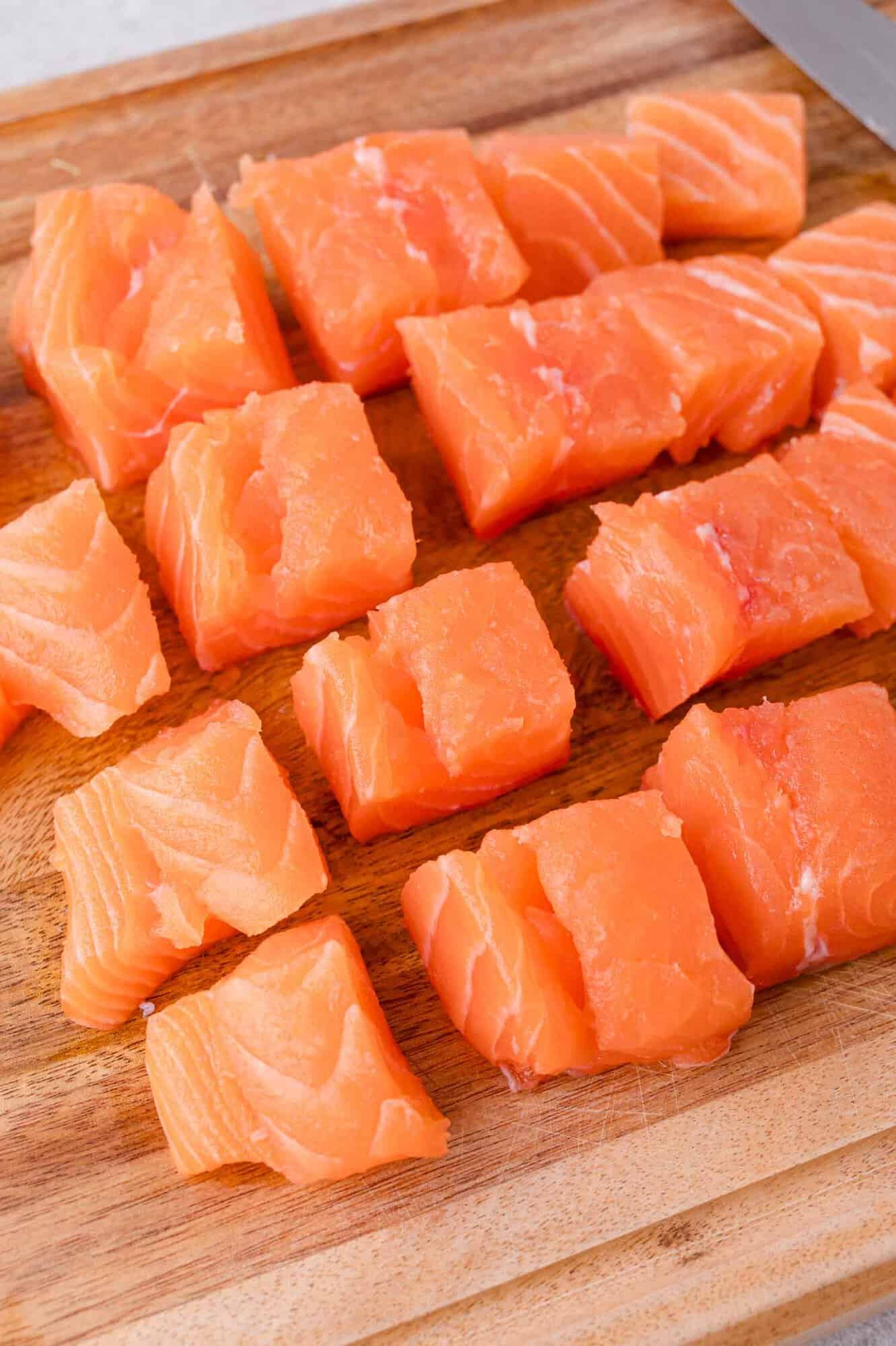 Cubes of raw salmon arranged on a wooden cutting board with a knife partially visible in the corner.