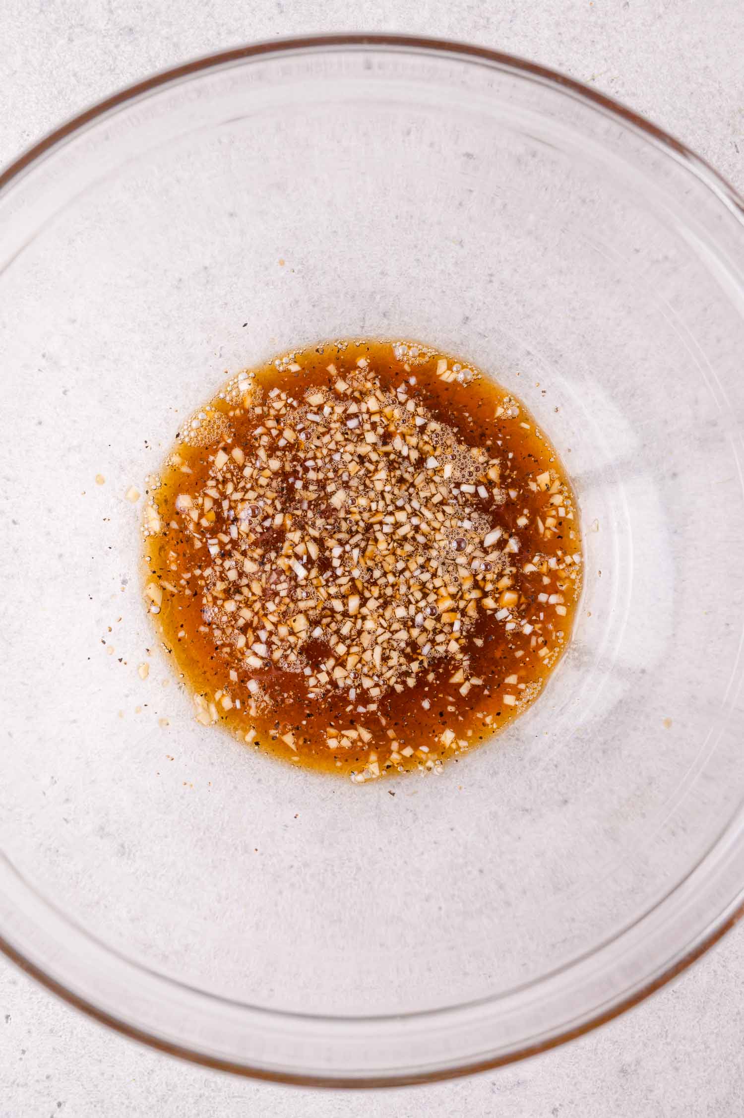 A clear glass bowl containing a mixture of brown liquid with visible minced garlic and spices on a light countertop.
