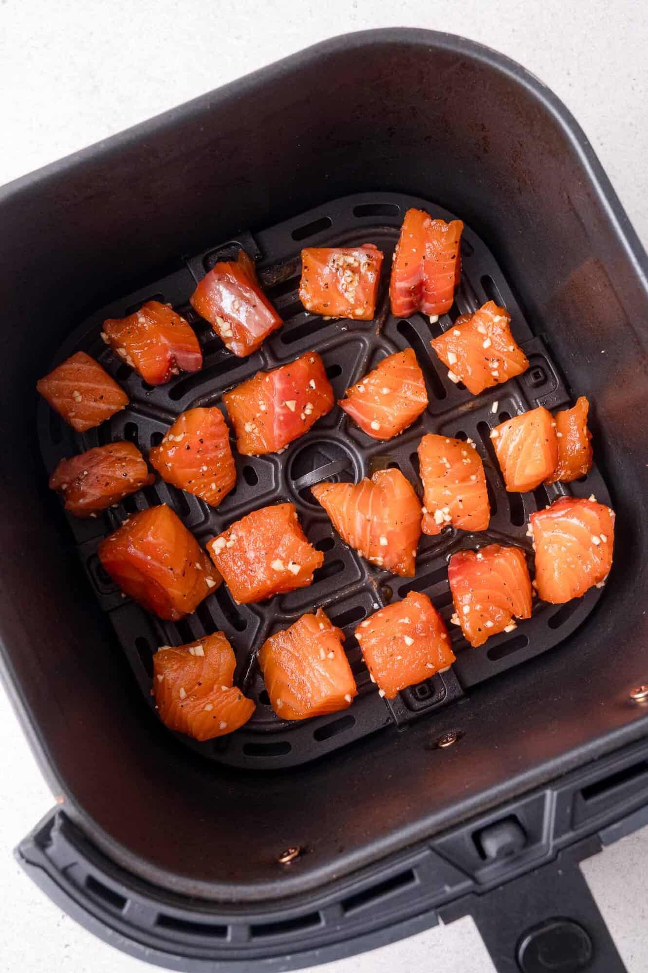 Cubed raw salmon pieces, seasoned with spices, are arranged in a single layer inside an air fryer basket on a light-colored countertop.
