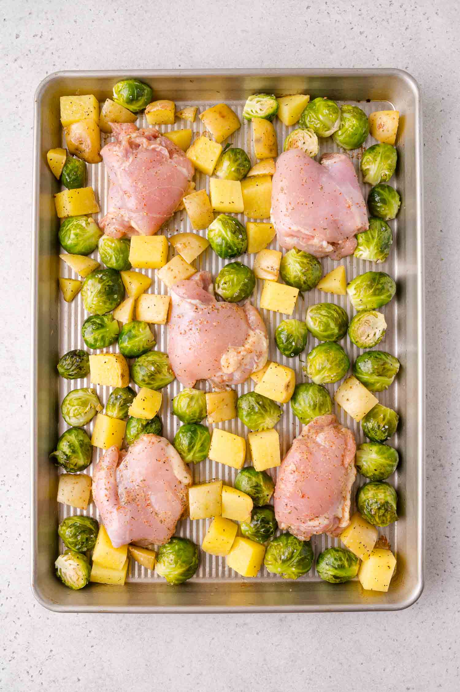 Raw chicken thighs, halved Brussels sprouts, and diced potatoes are evenly spaced on a metal baking sheet, ready for roasting.
