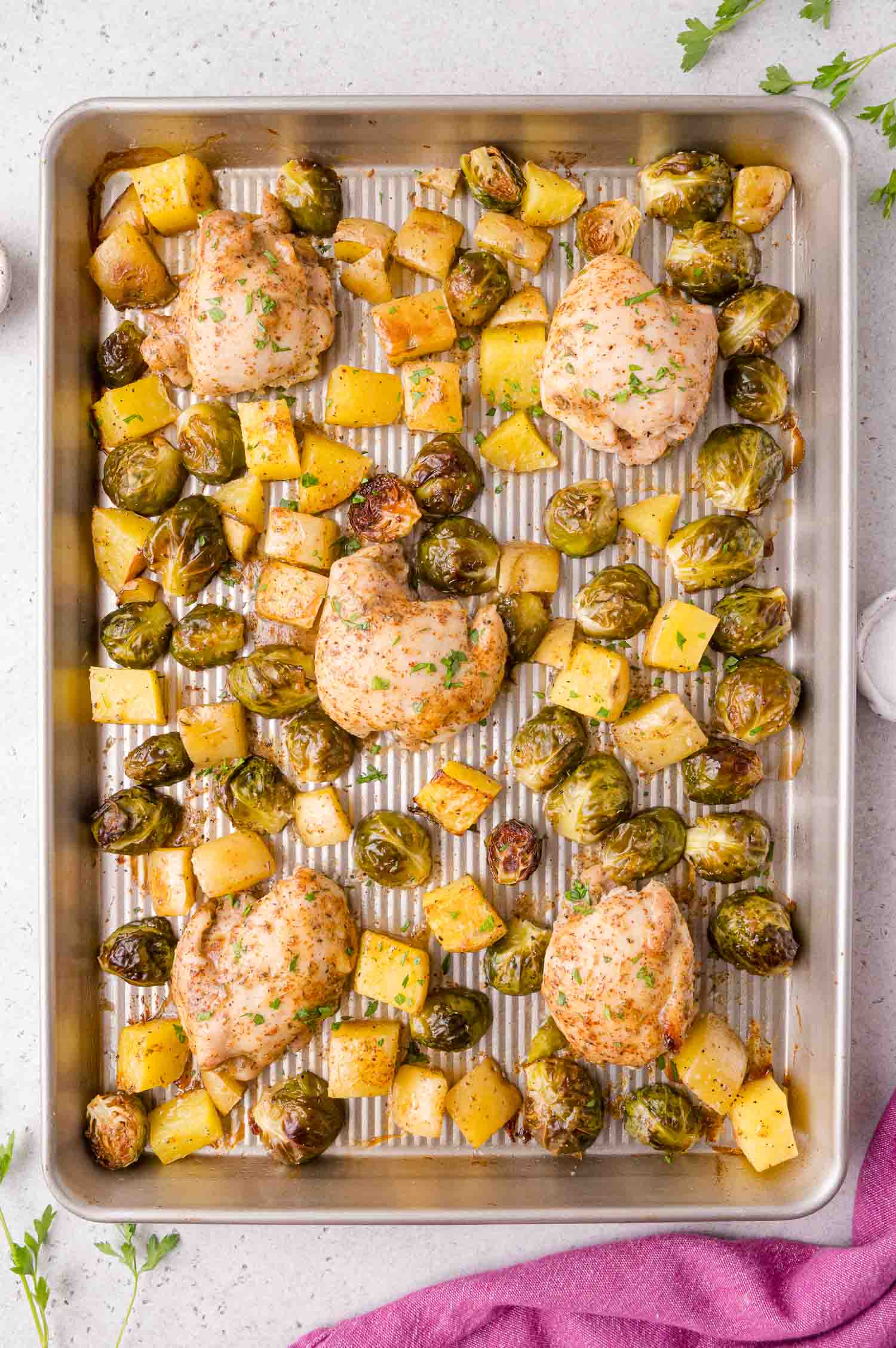 Sheet pan with baked chicken thighs, Brussels sprouts, and diced potatoes, seasoned and garnished with herbs, on a metal tray.
