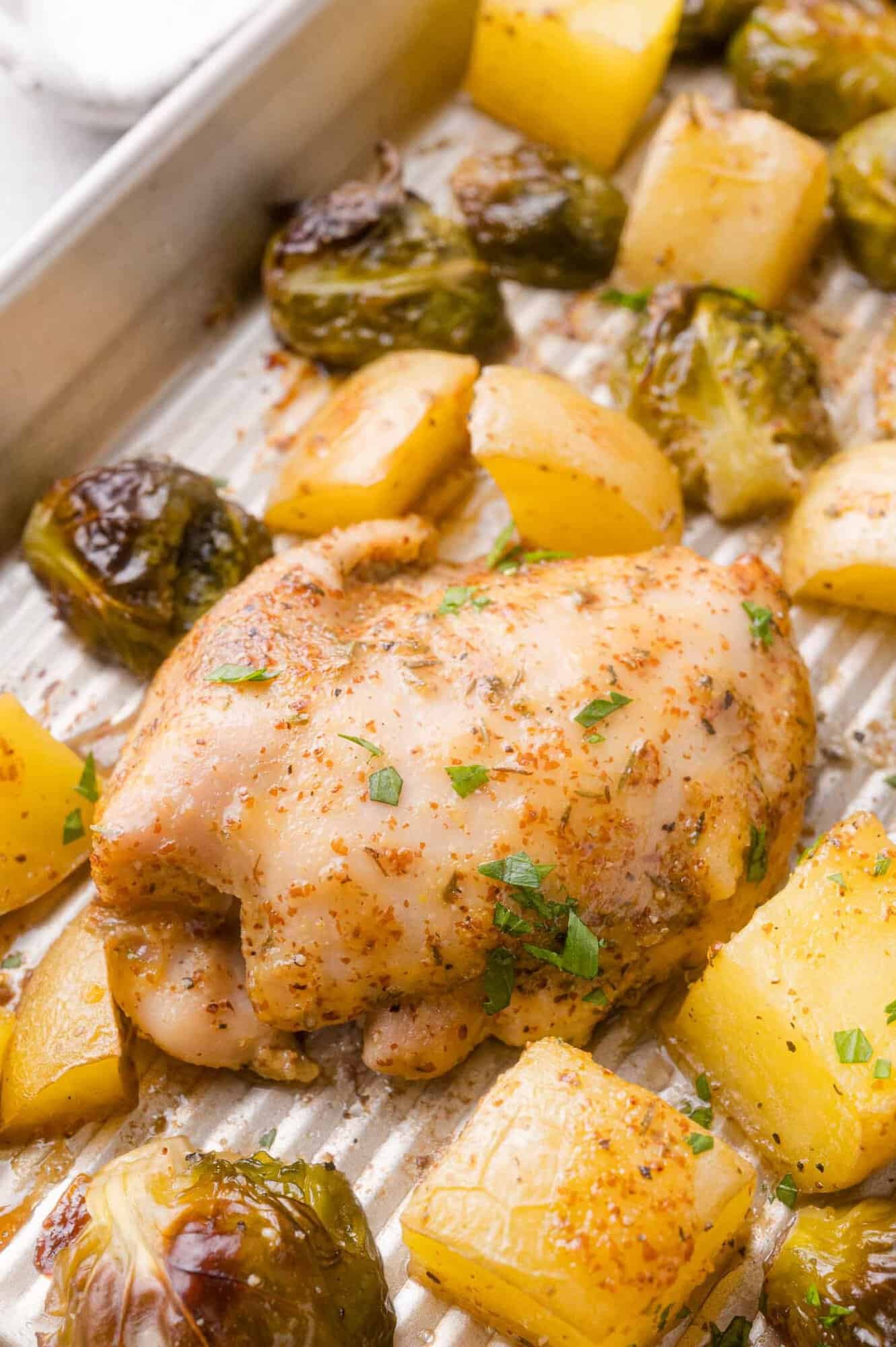 Baked chicken thigh with roasted brussels sprouts and potato chunks on a metal baking tray, garnished with herbs.