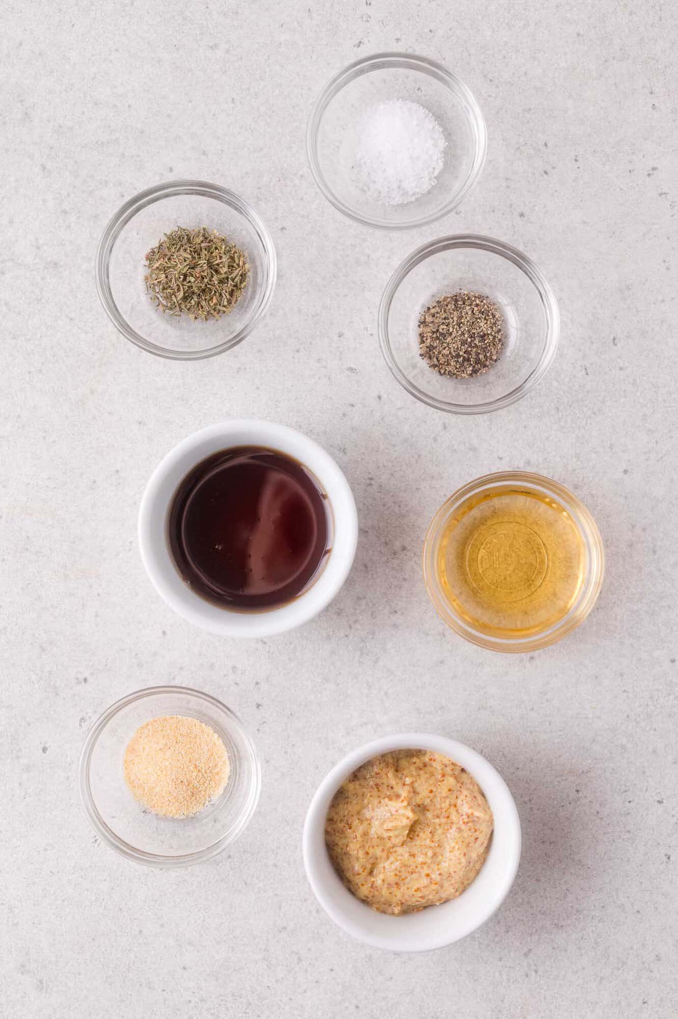 Seven small bowls containing salt, pepper, dried thyme, apple cider vinegar, maple syrup, garlic powder, and mustard are arranged on a light gray surface.