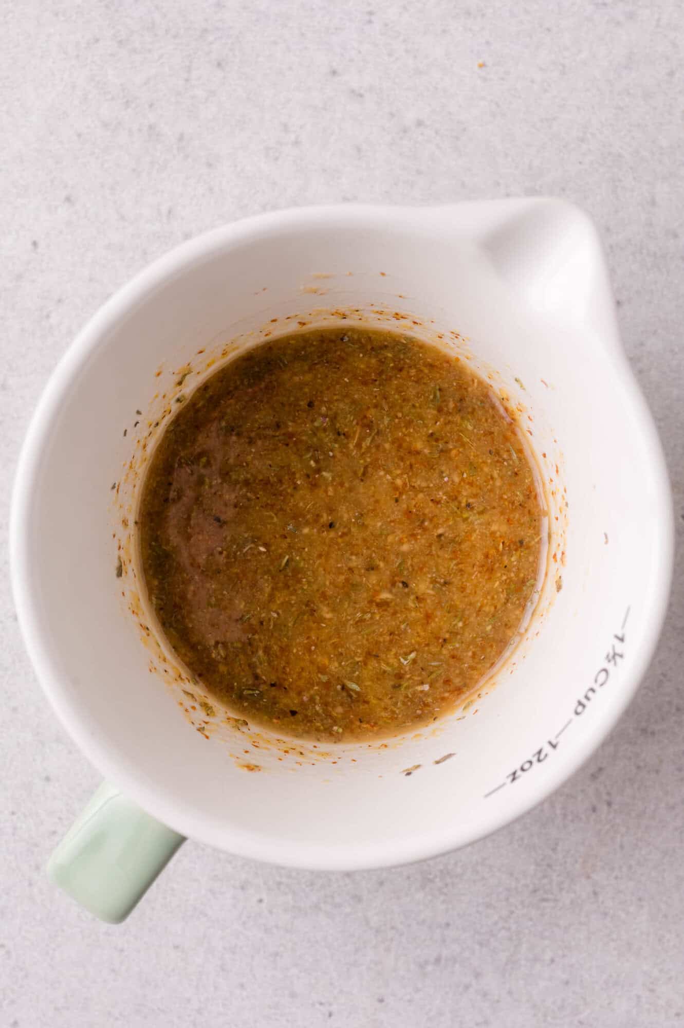 A white measuring cup containing a brown, mustard-based sauce mixture sits on a light gray surface.