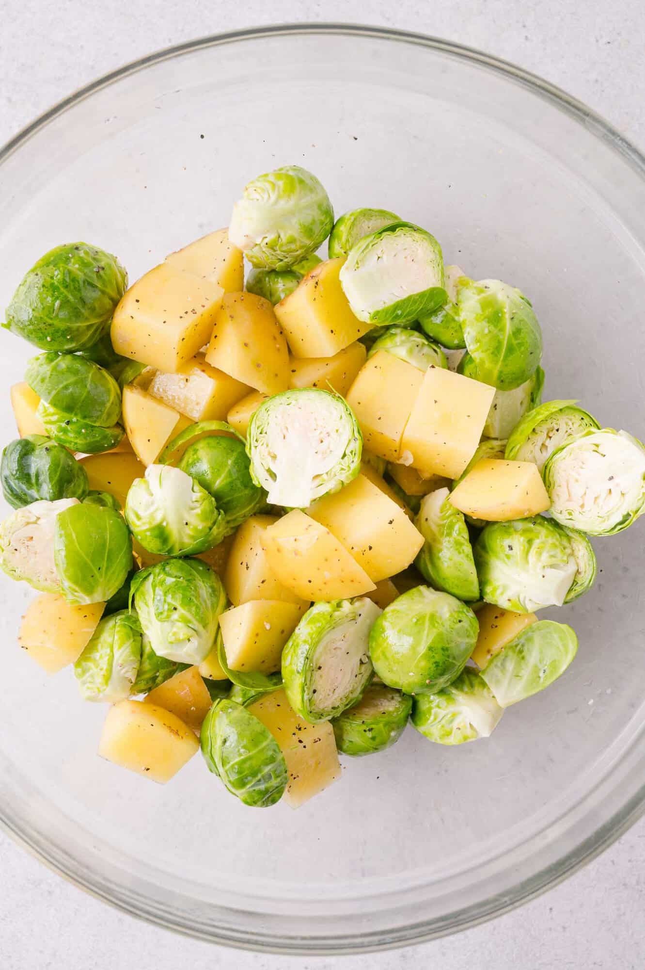 A glass bowl containing halved Brussels sprouts and cubed potatoes, lightly seasoned with salt and pepper.