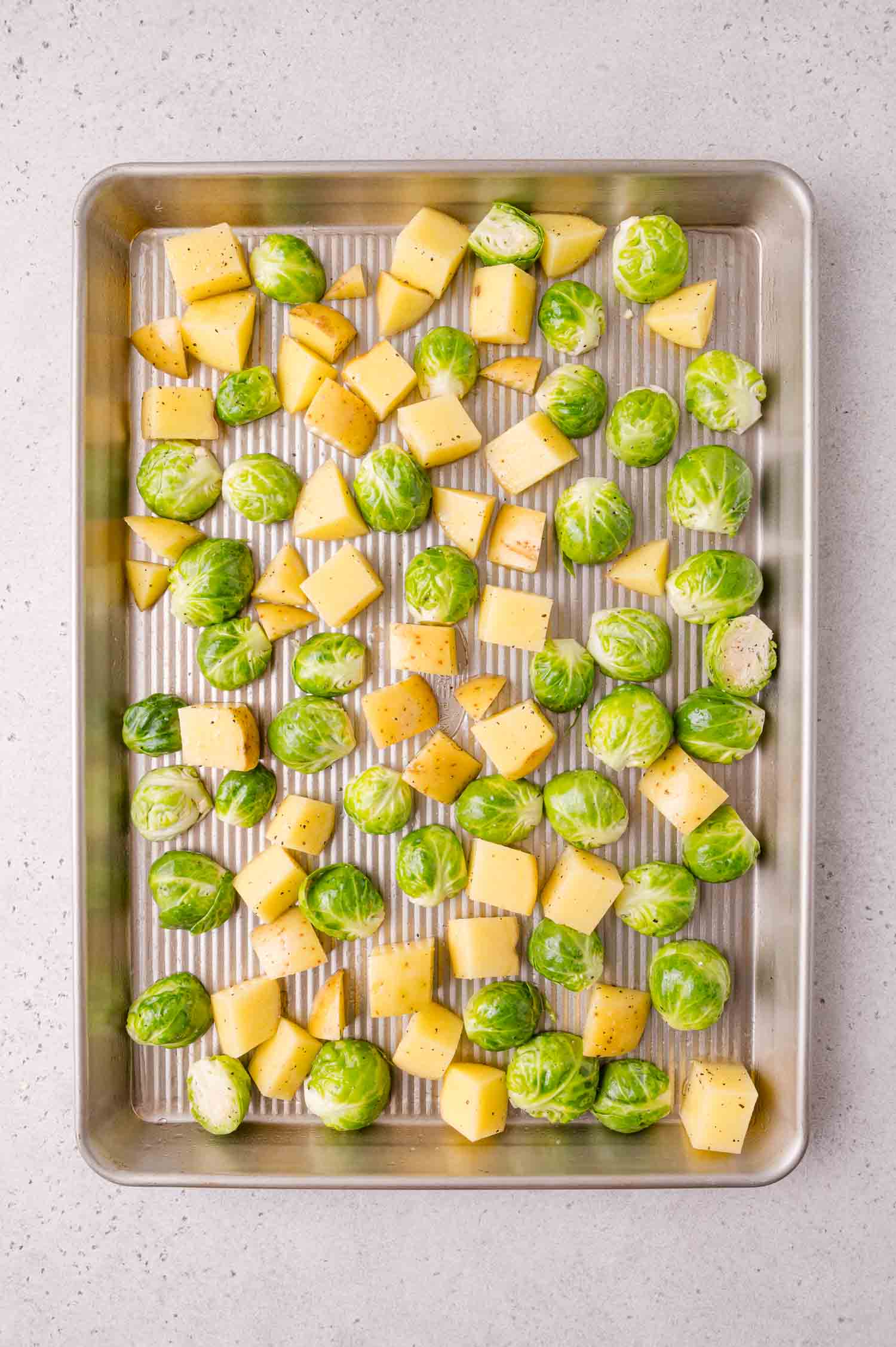 A baking sheet with raw Brussels sprouts and cubed potatoes, seasoned with salt and pepper, ready for roasting.