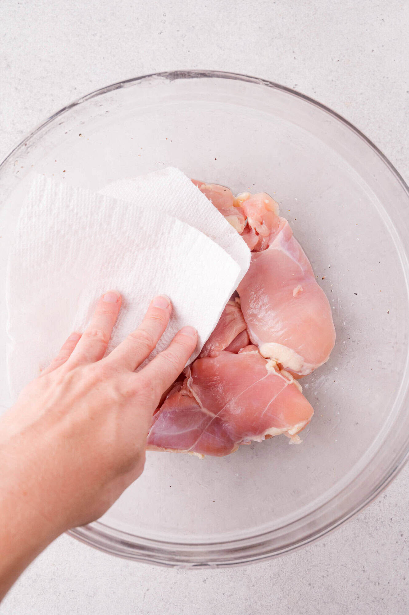 A hand uses a paper towel to pat raw chicken thighs dry in a clear glass bowl on a light-colored countertop.