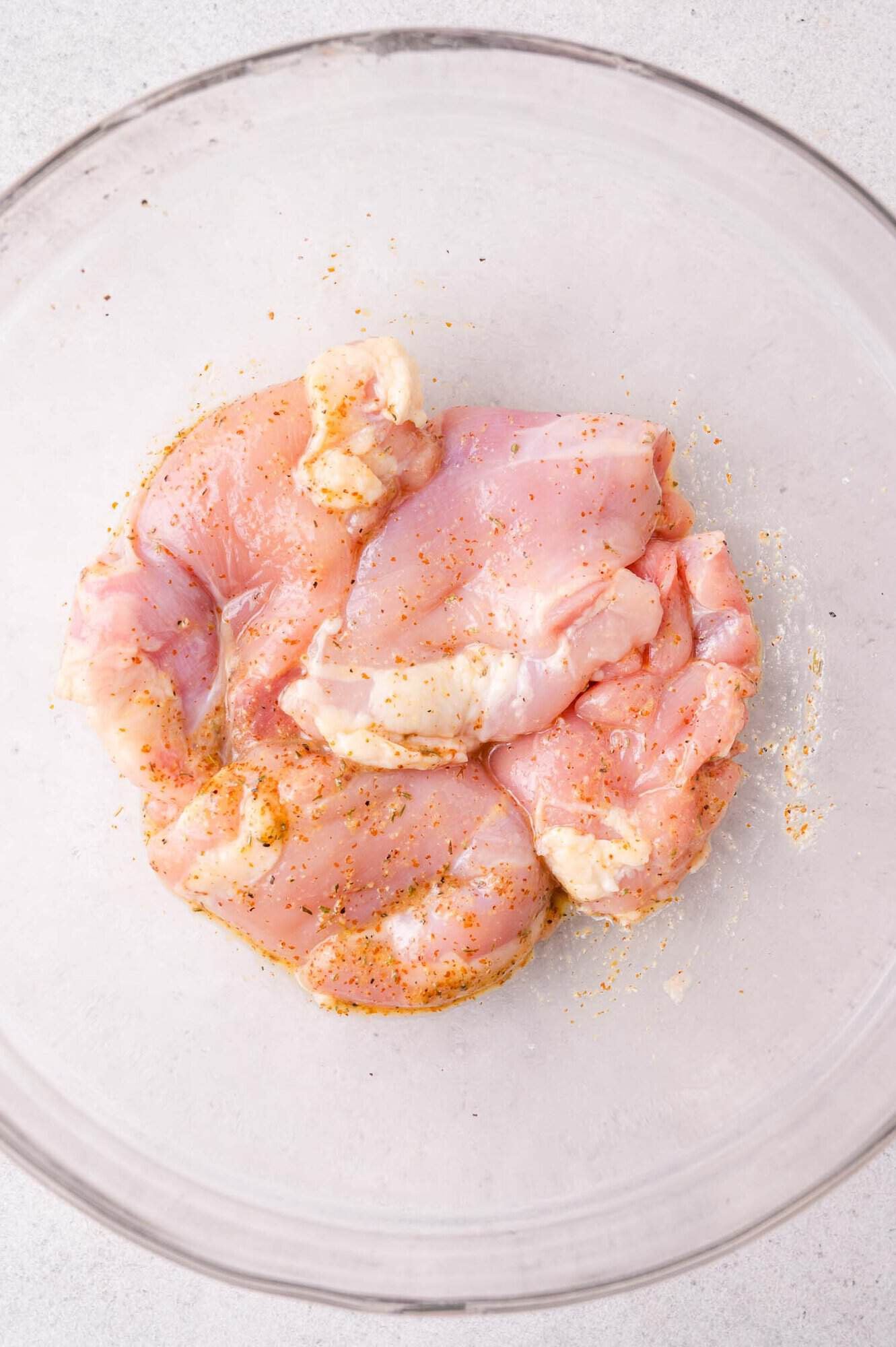 Raw, seasoned chicken pieces in a clear glass bowl on a light surface.